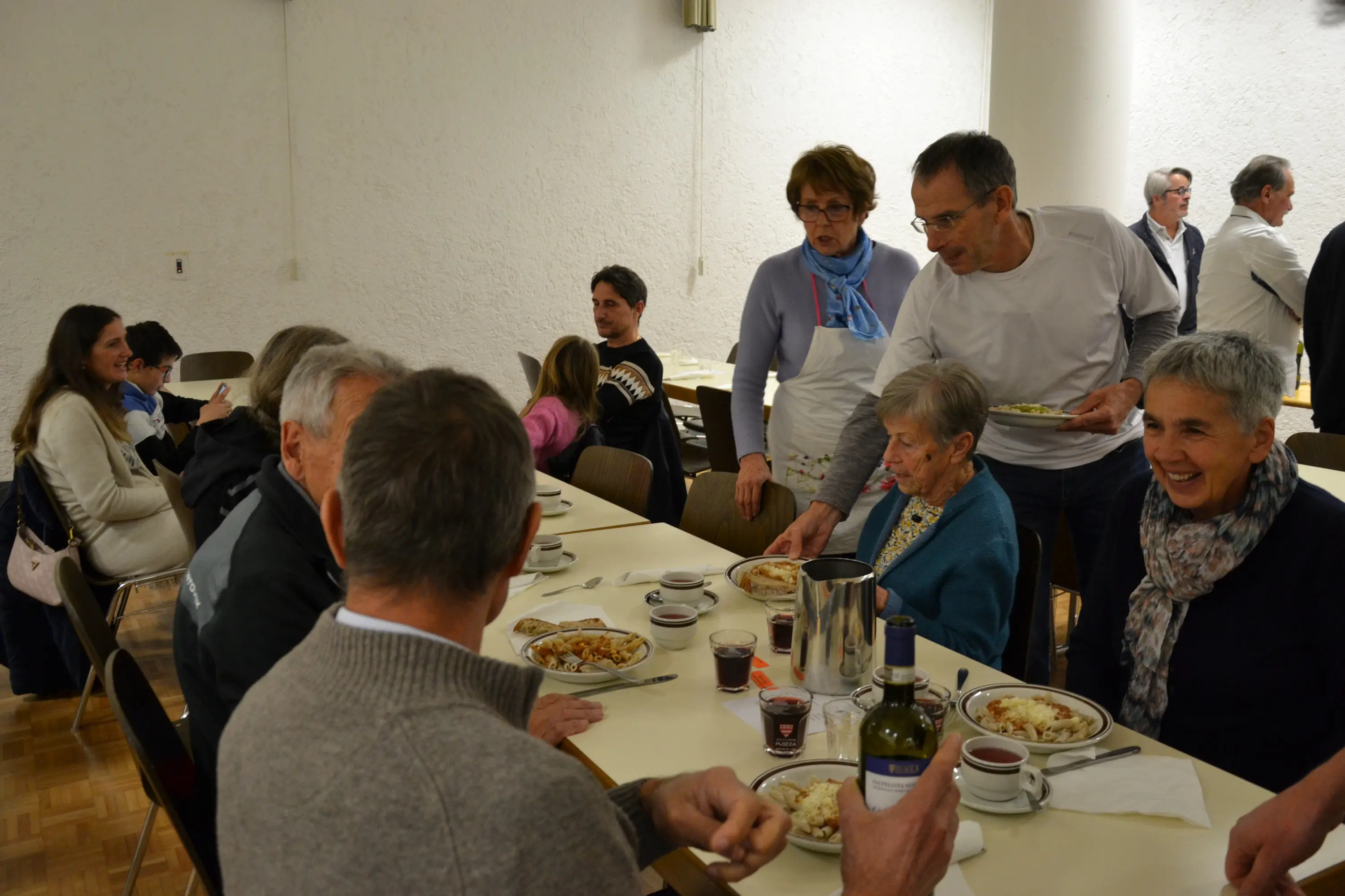 Una sala di ristorante con persone sedute a tavoli che condividono un pasto. Alcuni camerieri servono piatti e bevande. L'atmosfera è conviviale e informale.