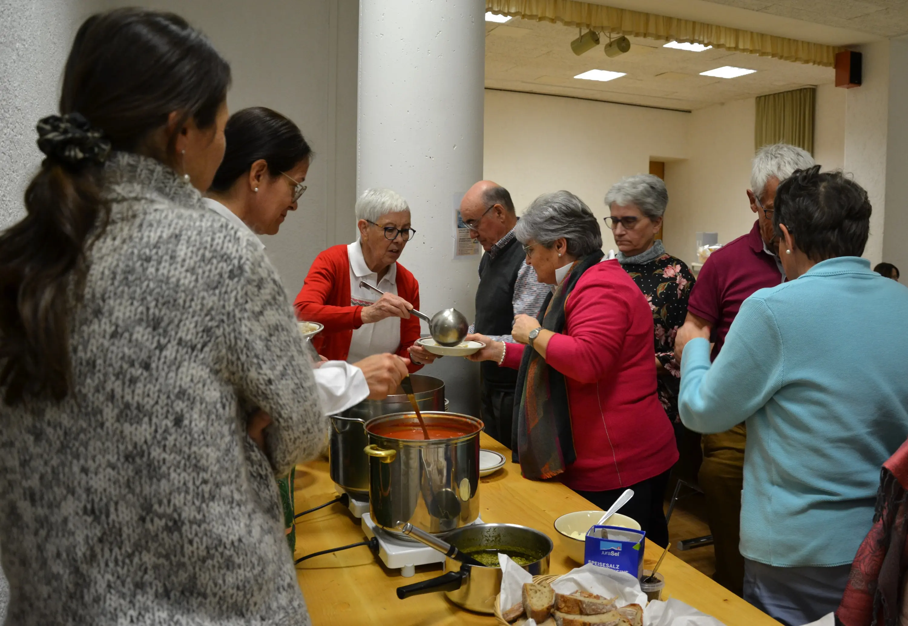 Un gruppo di persone si riunisce attorno a un tavolo, servendo e condividendo cibo da pentole. Alcuni stanno ricevendo porzioni, mentre altri chiacchierano. I partecipanti indossano abiti caldi e l'ambiente appare accogl