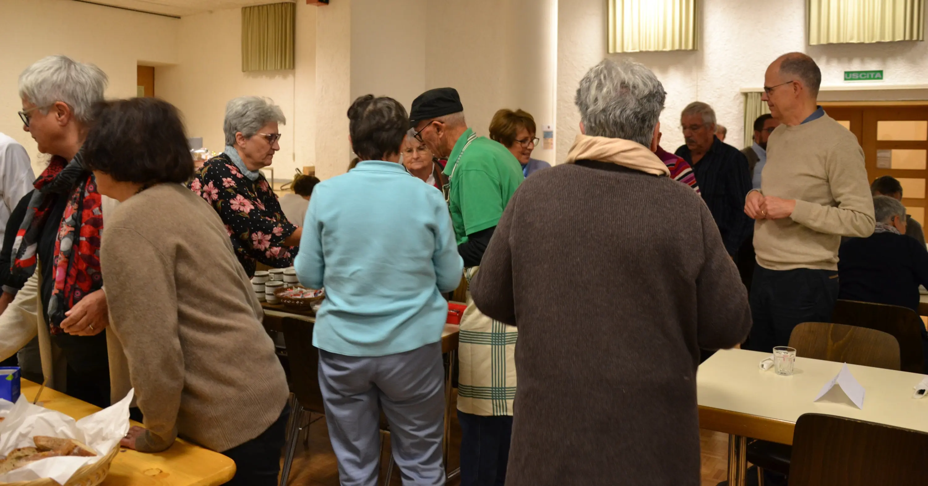 Persone di diverse età si radunano in una sala per partecipare a un evento sociale. Alcuni stanno servendo cibo mentre altri chiacchierano tra loro.