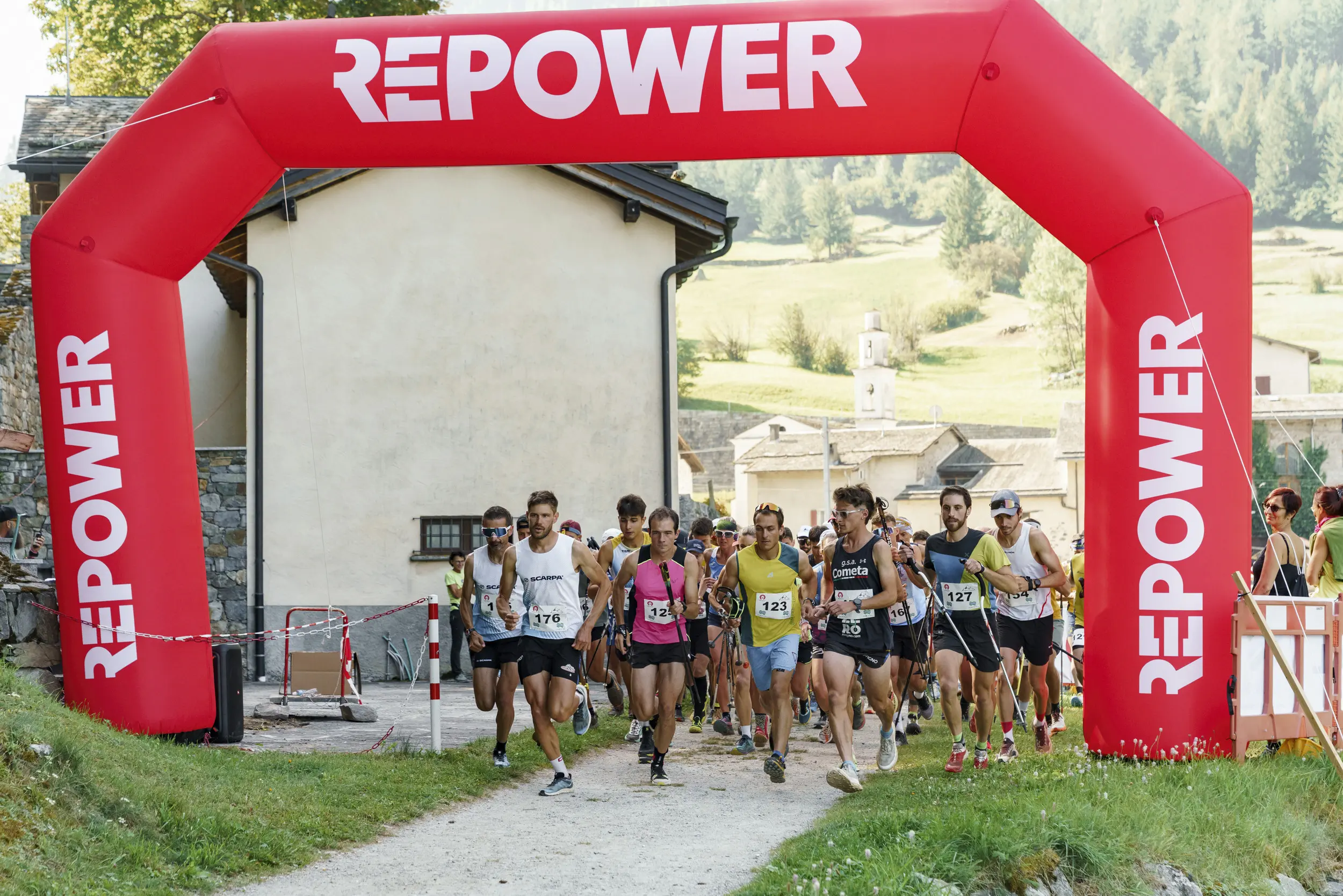 Partecipanti che attraversano un arco gonfiabile rosso con la scritta "REPOWER" all'inizio di una corsa, circondati da un paesaggio montano.