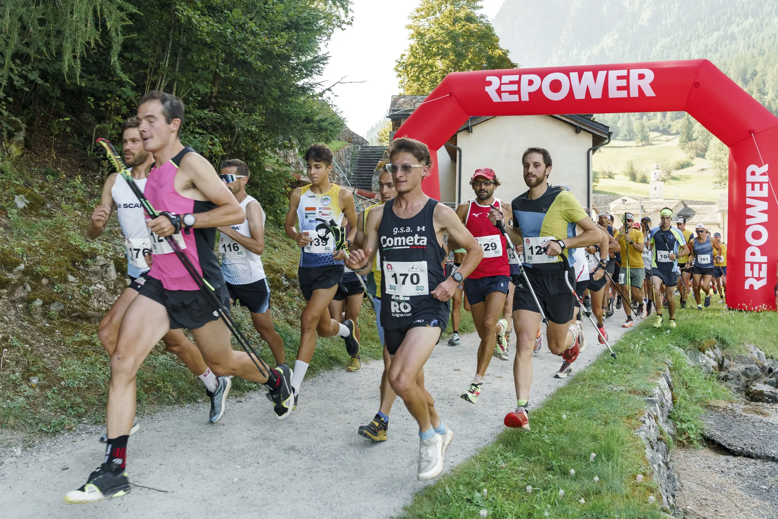 Atleti in gara partecipano a una corsa su un sentiero, passando sotto un arco gonfiabile con la scritta "REPOWER". Sullo sfondo si vedono alberi e montagne.