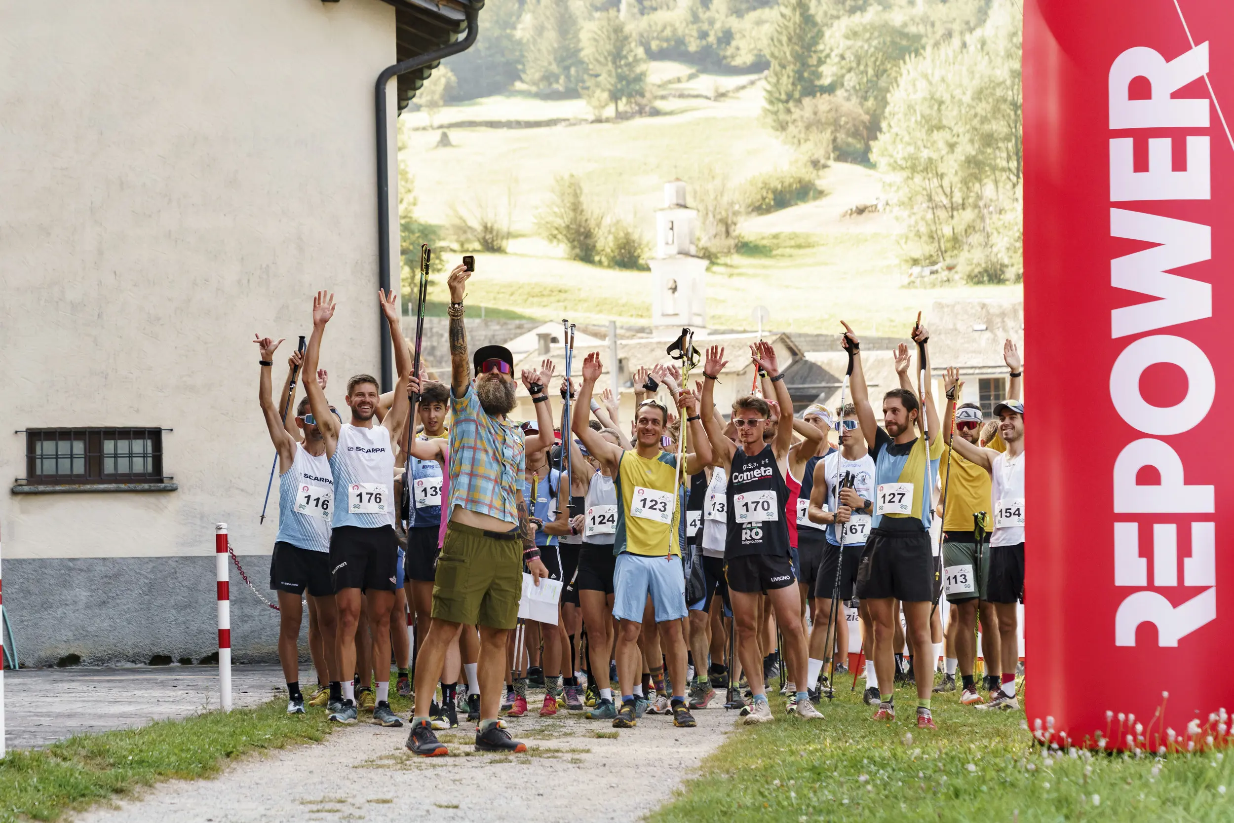 Partecipanti di una corsa si radunano sotto un arco rosso con la scritta "REPOWER". Alcuni alzano le mani celebrando, mentre altri sono pronti a partire. Sullo sfondo si notano colline verdi.
