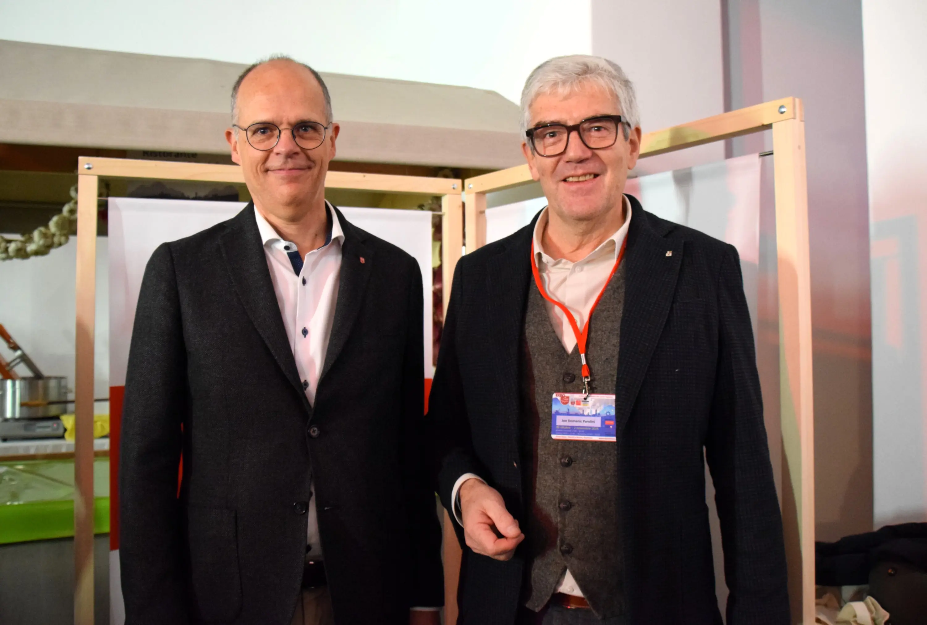 Due uomini sorridenti posano insieme, indossando abiti formali. Si trovano in un ambiente commerciale, con attrezzature e poster sullo sfondo.