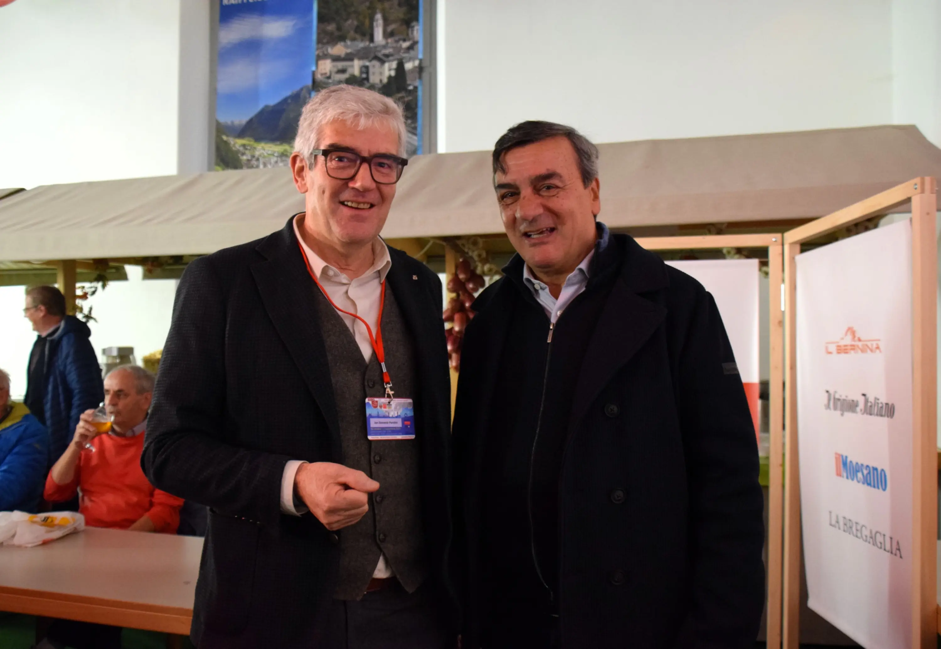 Due uomini sorridenti posano insieme in un ambiente accogliente, con una tenda sullo sfondo. Uno indossa occhiali e un cardigan, mentre l'altro ha un cappotto scuro. Ci sono persone sedute dietro di loro, e un cartello sulla des