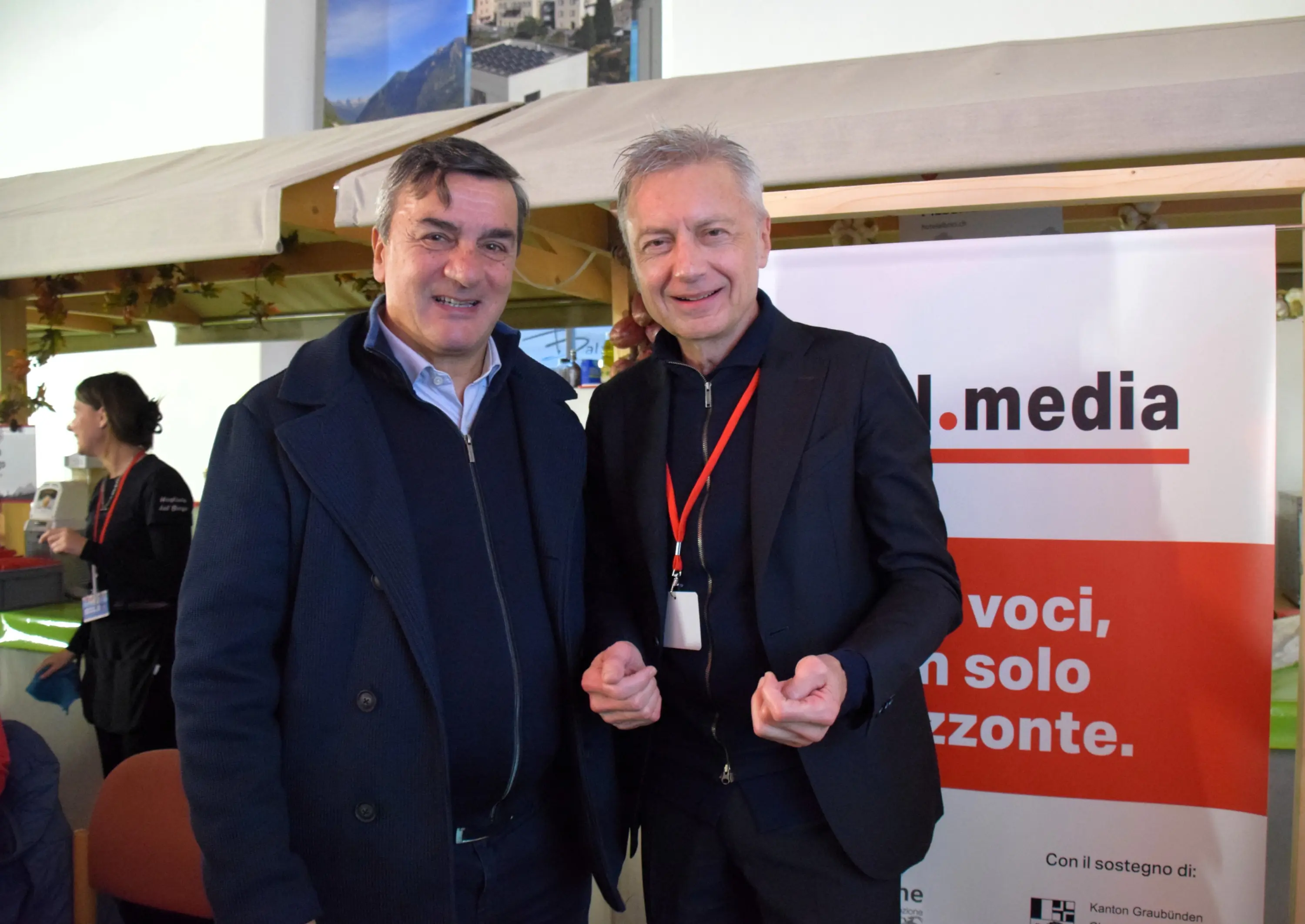 Due uomini sorridenti posano fianco a fianco, indossando giacche. Sullo sfondo è visibile un cartello con il logo di un'agenzia media.