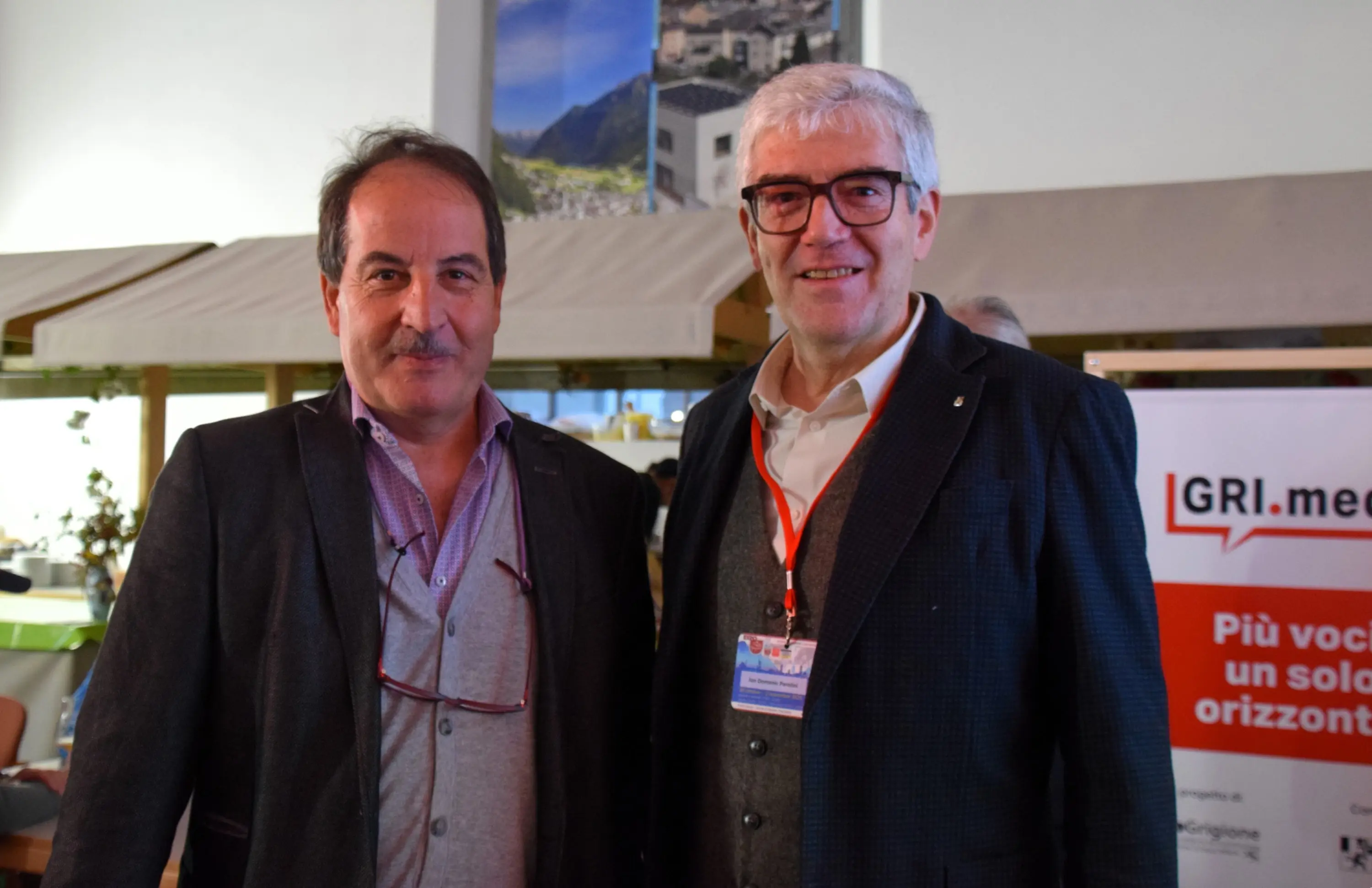 Due uomini sorridenti si trovano in un ambiente accogliente. Indossano abiti formali e occhiali. Dietro di loro si intravede un banner con la scritta "GRI.med" e un paesaggio montano.