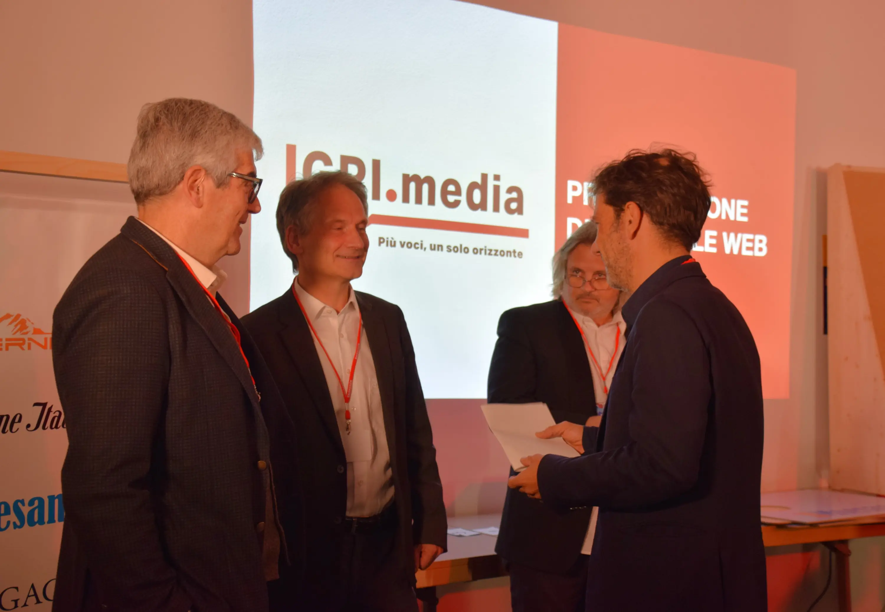 Un gruppo di uomini in abiti formali interagisce durante un evento, con un logo di ICBI.media sullo sfondo. Uno degli uomini sta leggendo un documento, mentre gli altri ascoltano attentamente.