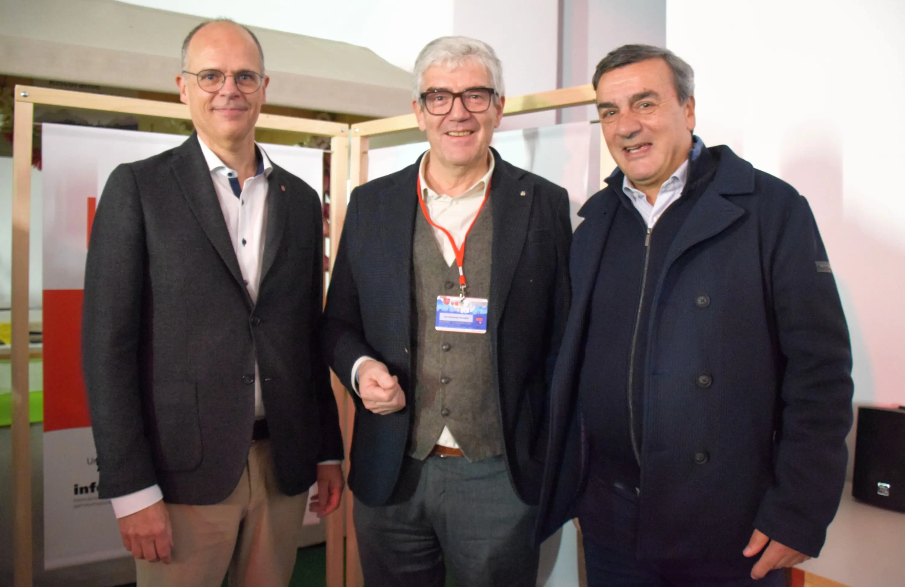 Tre uomini sorridenti posano insieme in un ambiente moderno, con una struttura di legno sullo sfondo. Indossano abbigliamento elegante e casual, mentre uno di loro tiene una scheda identificativa al collo.