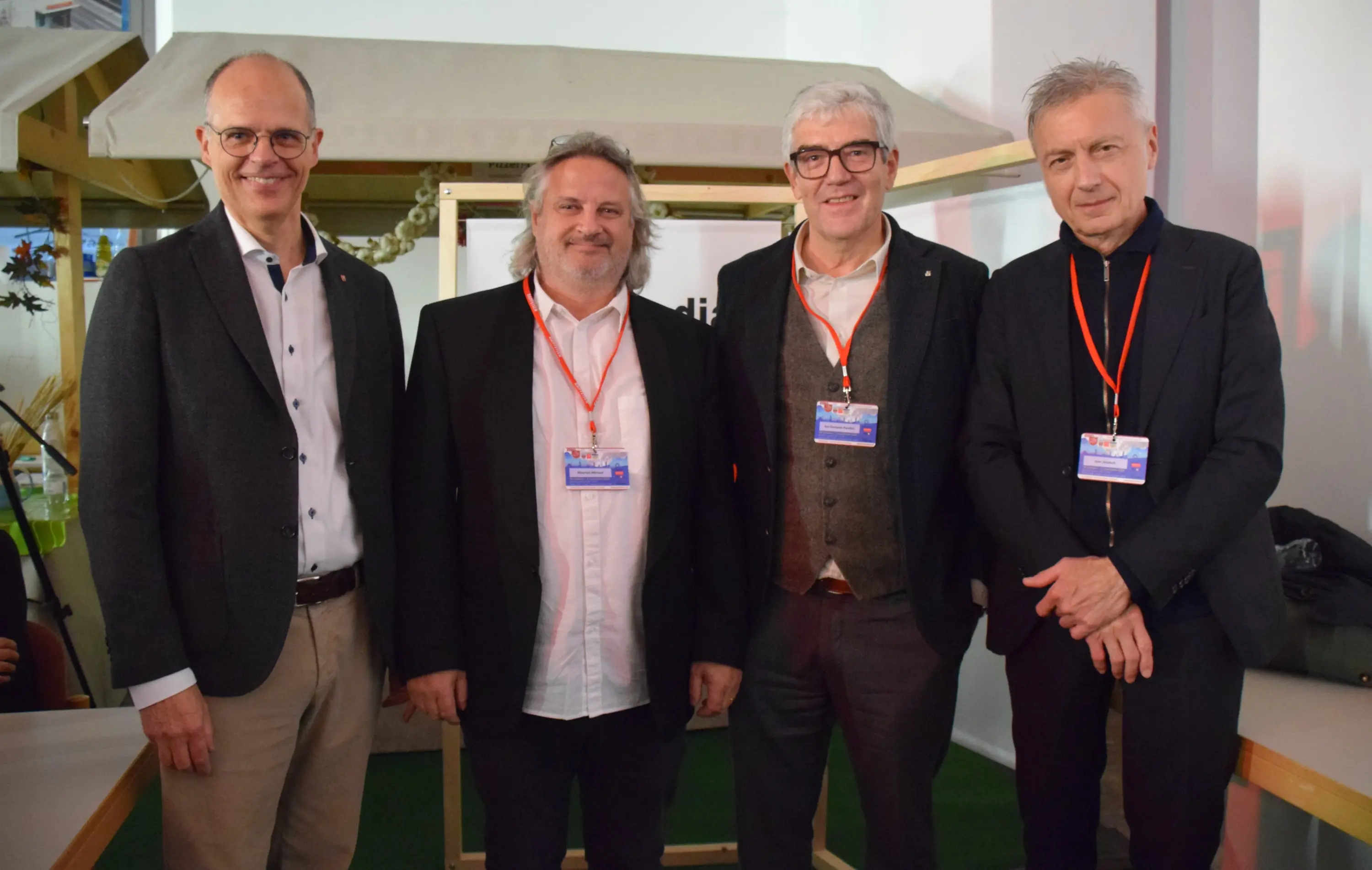 Quattro uomini in abbigliamento formale sorridono e posano insieme, mentre indossano dei badge identificativi. Sfondo neutro con dettagli indistinti.