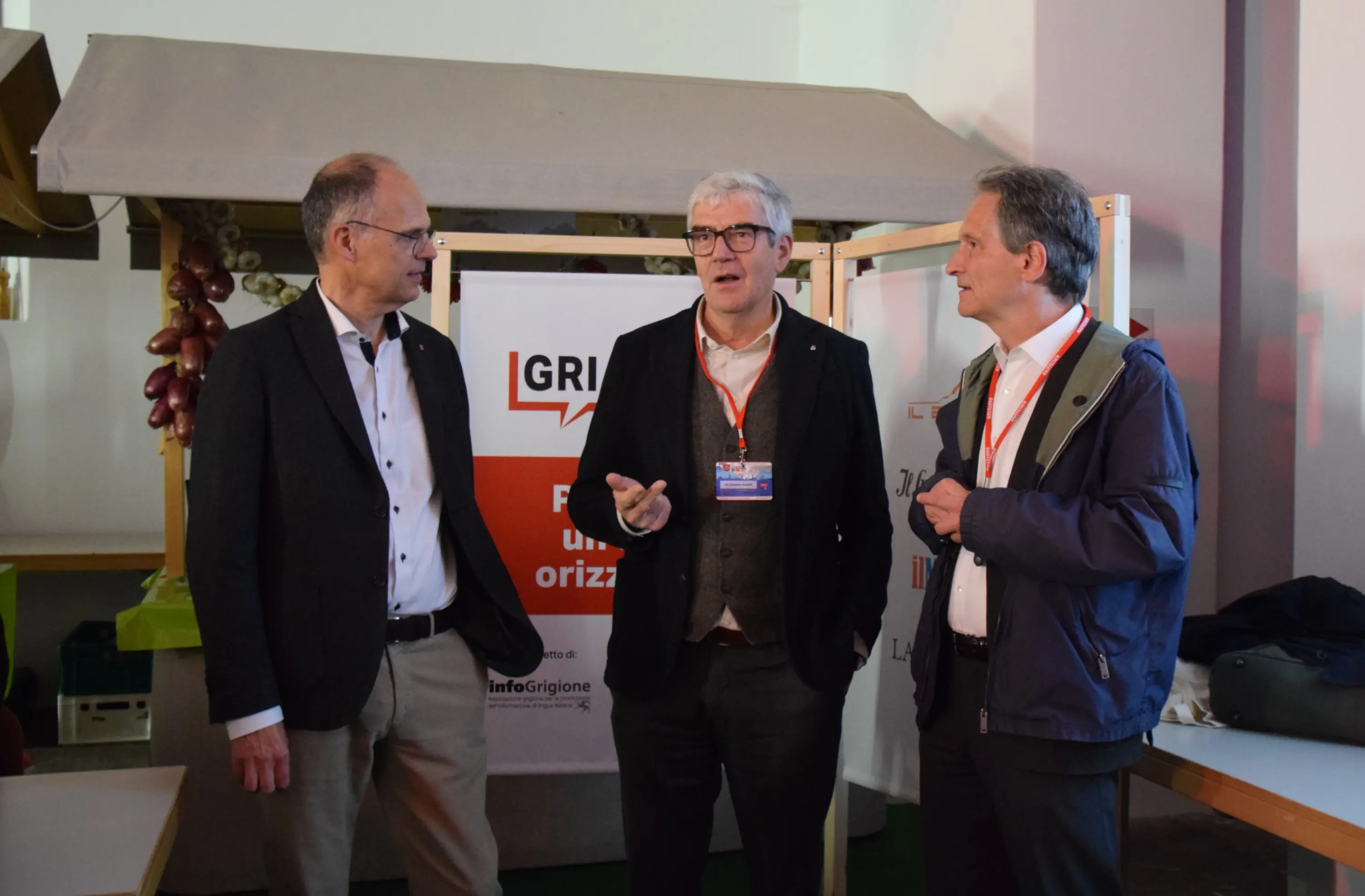 Tre uomini in abiti formali discutono davanti a uno sfondo con un banner che promuove un evento. Due di loro indossano giacche e uno ha occhiali. L'atmosfera è cordiale e di scambio.