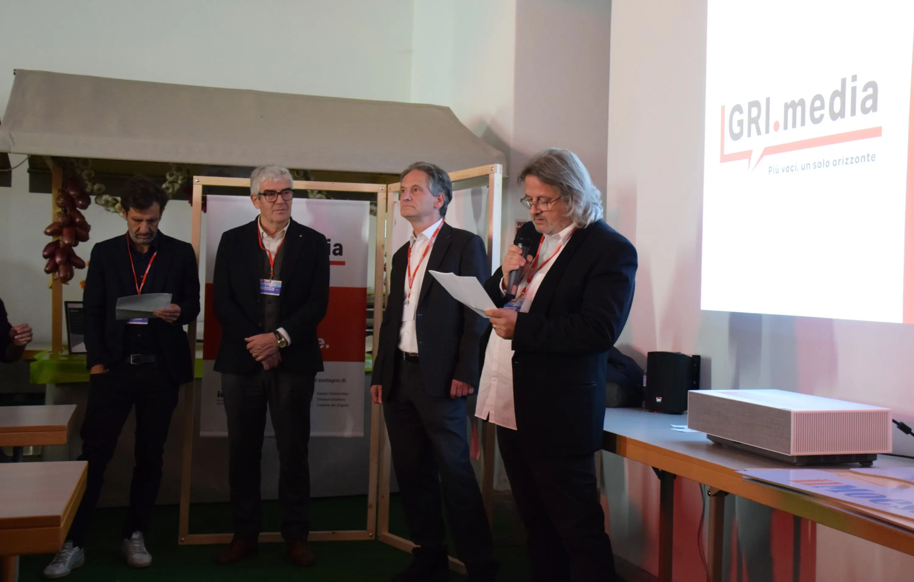 Un gruppo di cinque uomini sta partecipando a un evento, alcuni leggono documenti mentre altri ascoltano. Sullo sfondo, un grande schermo mostra il logo di GRI.media. L'atmosfera è di attenzione e professionalità.