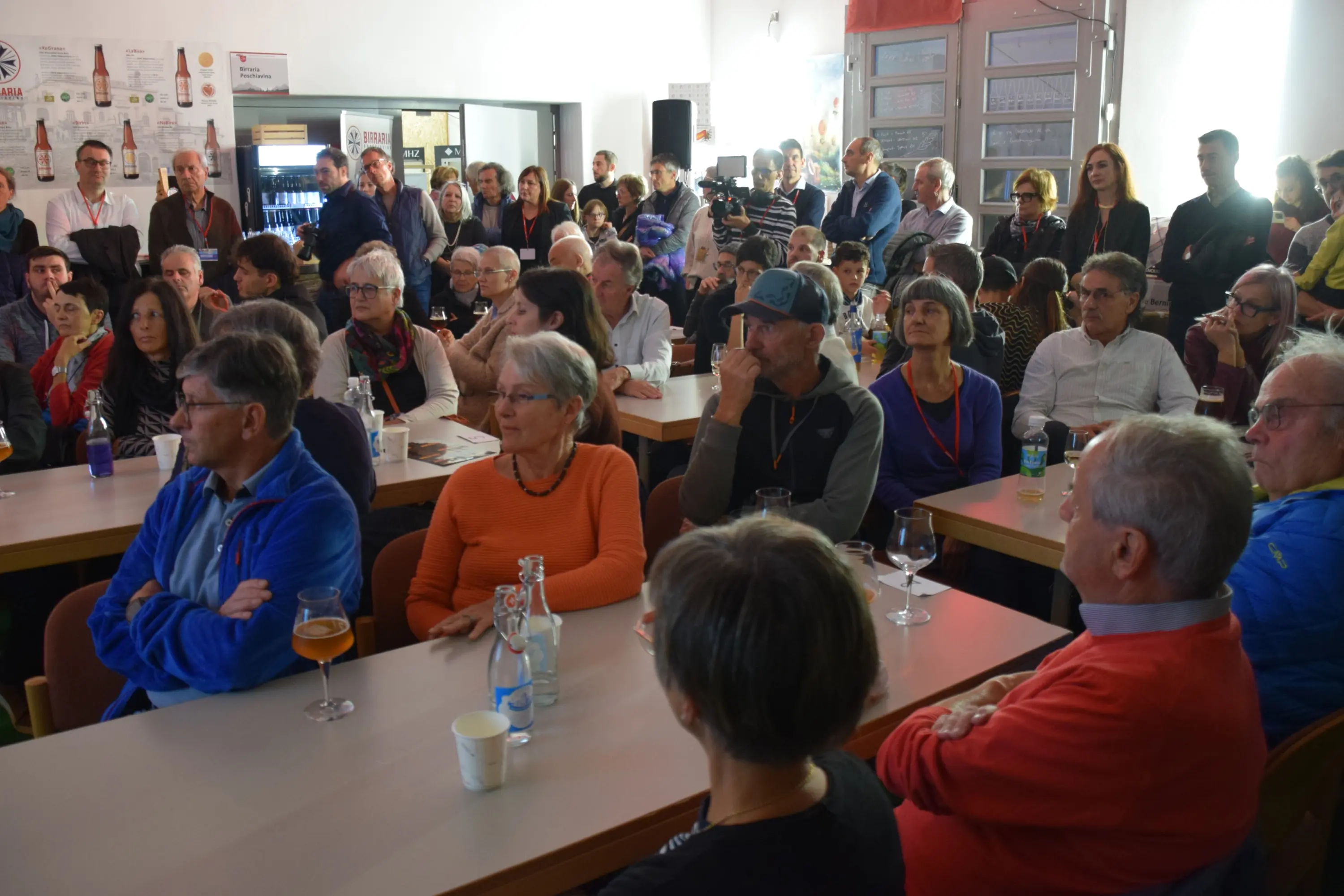 Una sala affollata con persone sedute a tavoli, molte stanno ascoltando attentamente un relatore. Ci sono bevande sui tavoli e un'atmosfera di interesse e coinvolgimento.