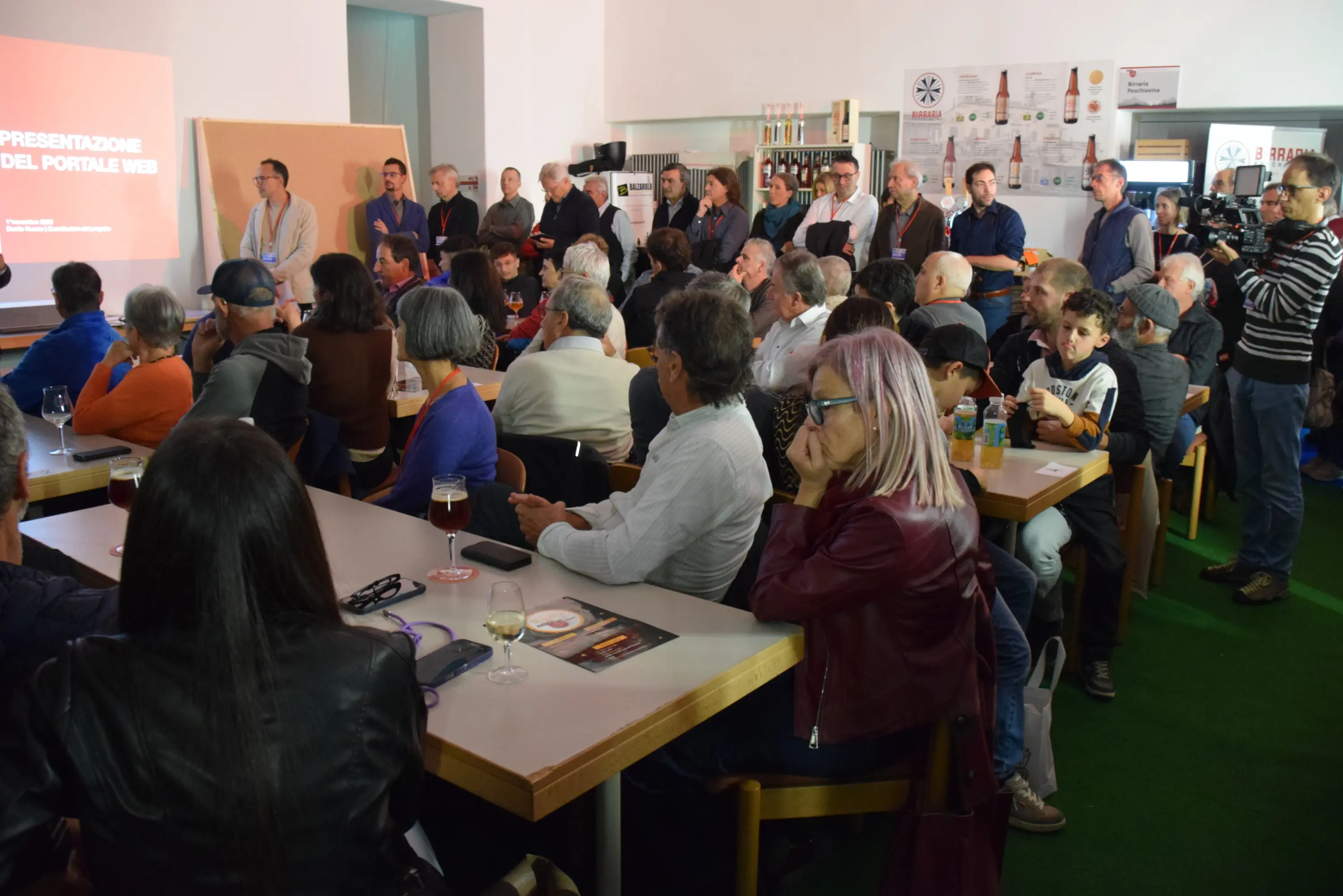 Un gruppo numeroso di persone ascolta una presentazione in una sala, con alcuni davanti a un palco e altri seduti ai tavoli. Il pubblico è composto da adulti e bambini, mentre sul tavolo si possono vedere bevande e cibo.