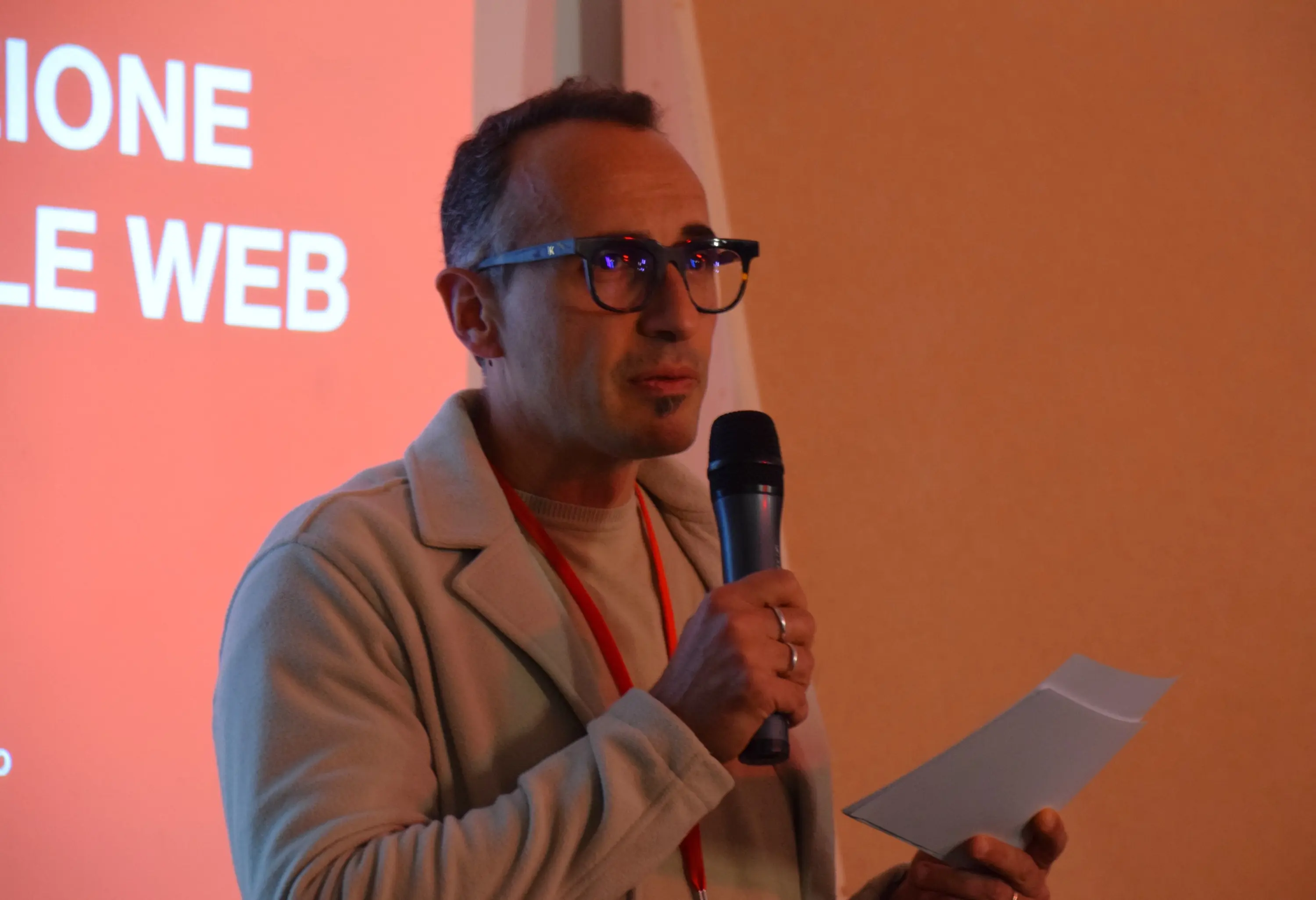 Un uomo in piedi di fronte a uno schermo con un testo rosso. Tiene un microfono in una mano e un foglio nell'altra. Indossa occhiali e un maglione chiaro.