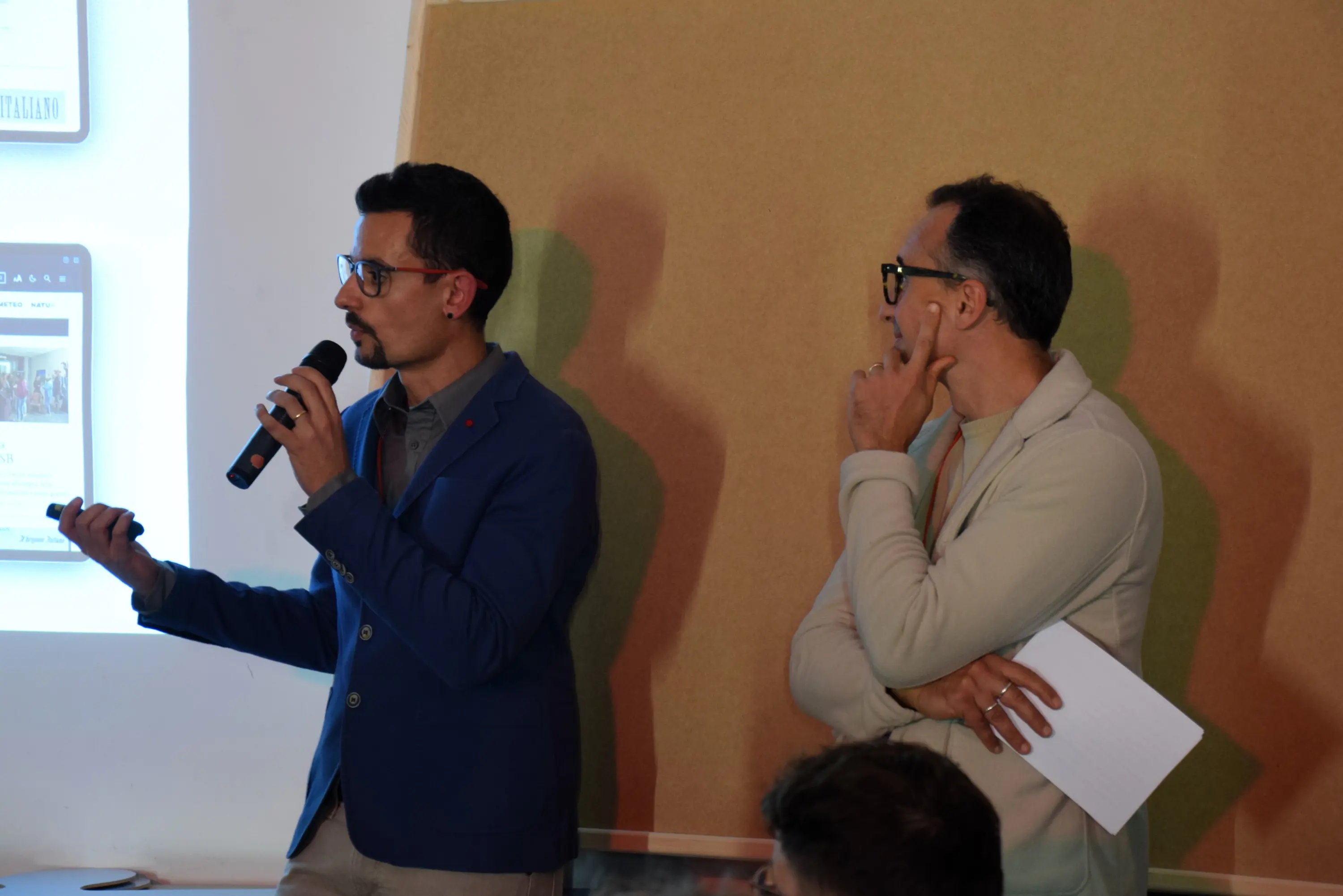 Due uomini in un contesto di presentazione. Il primo, con occhiali e giacca blu, parla al microfono mentre indica uno schermo. Il secondo, con occhiali e maglione chiaro, ascolta attentamente.