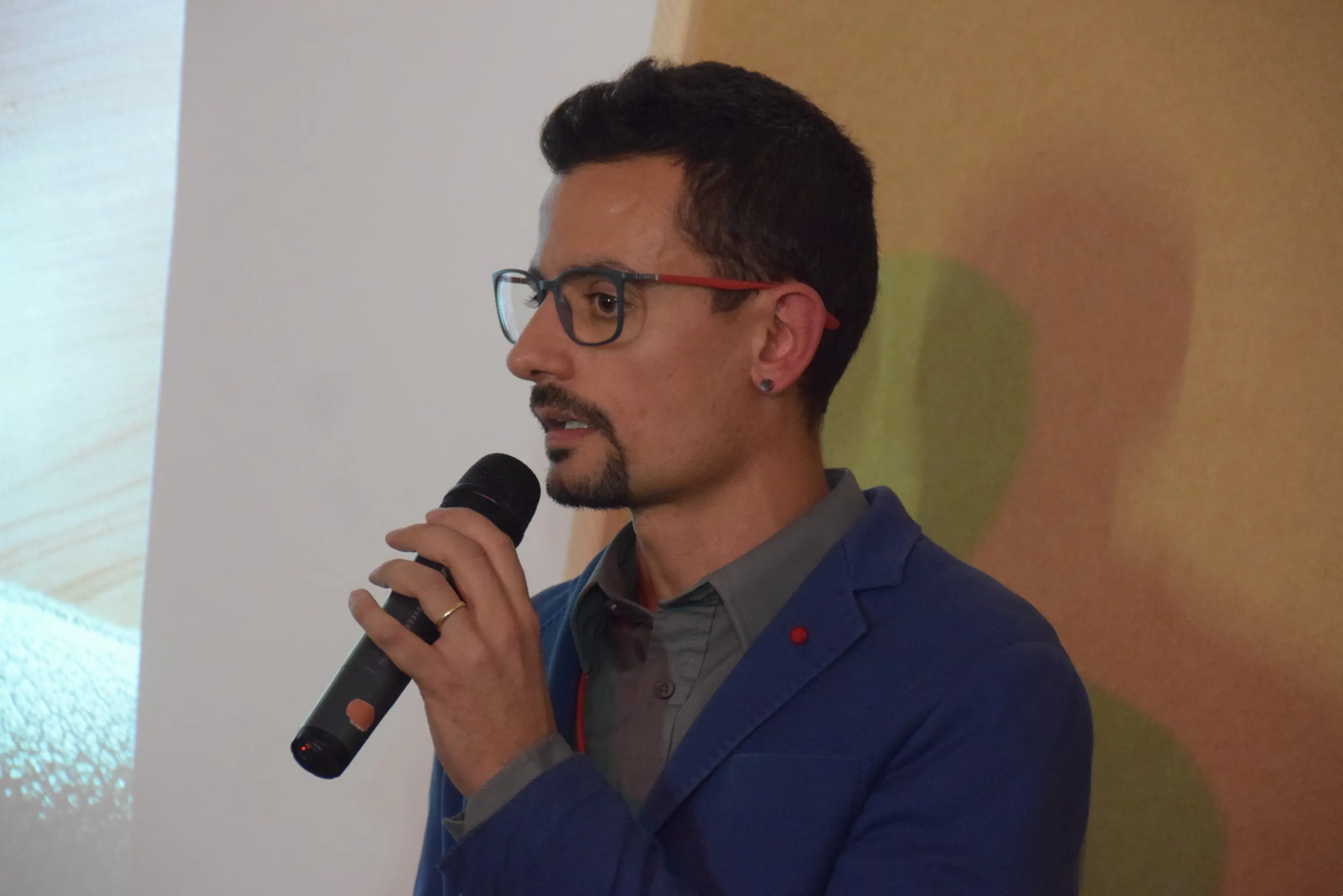 Un uomo con occhiali e una giacca blu parla al microfono, mentre sembra essere impegnato in una presentazione.