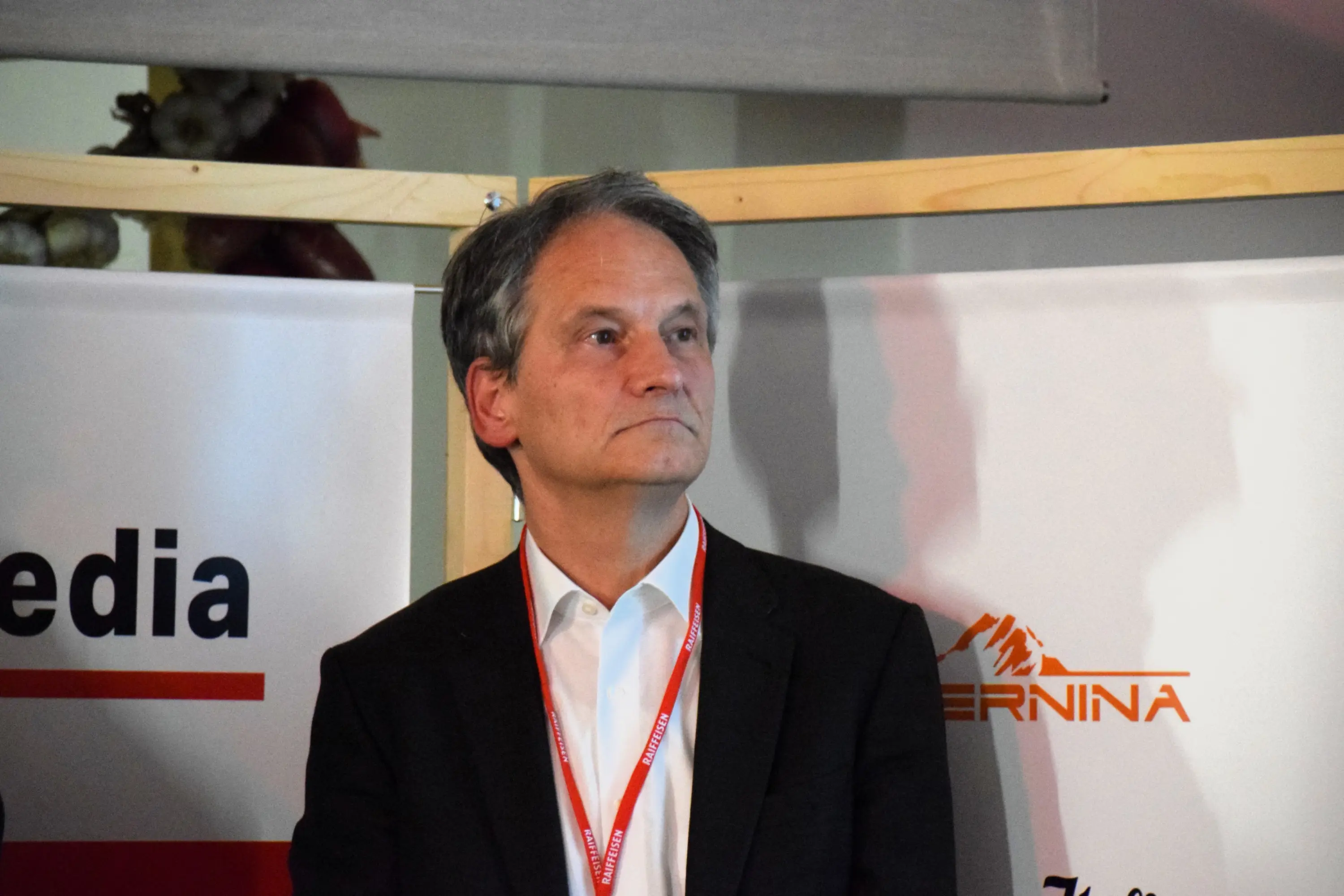 Uomo in abito scuro con camicia bianca, mentre osserva con espressione seria, di fronte a un pannello che riporta il logo di un evento.