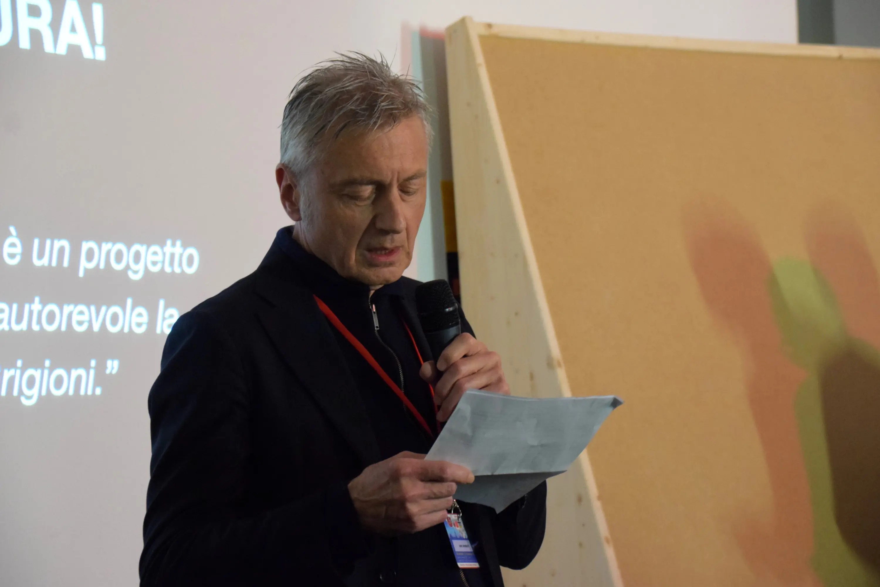 Uomo anziano in giacca nera che legge un testo con un microfono in mano. Sullo sfondo, una presentazione con un messaggio visibile.