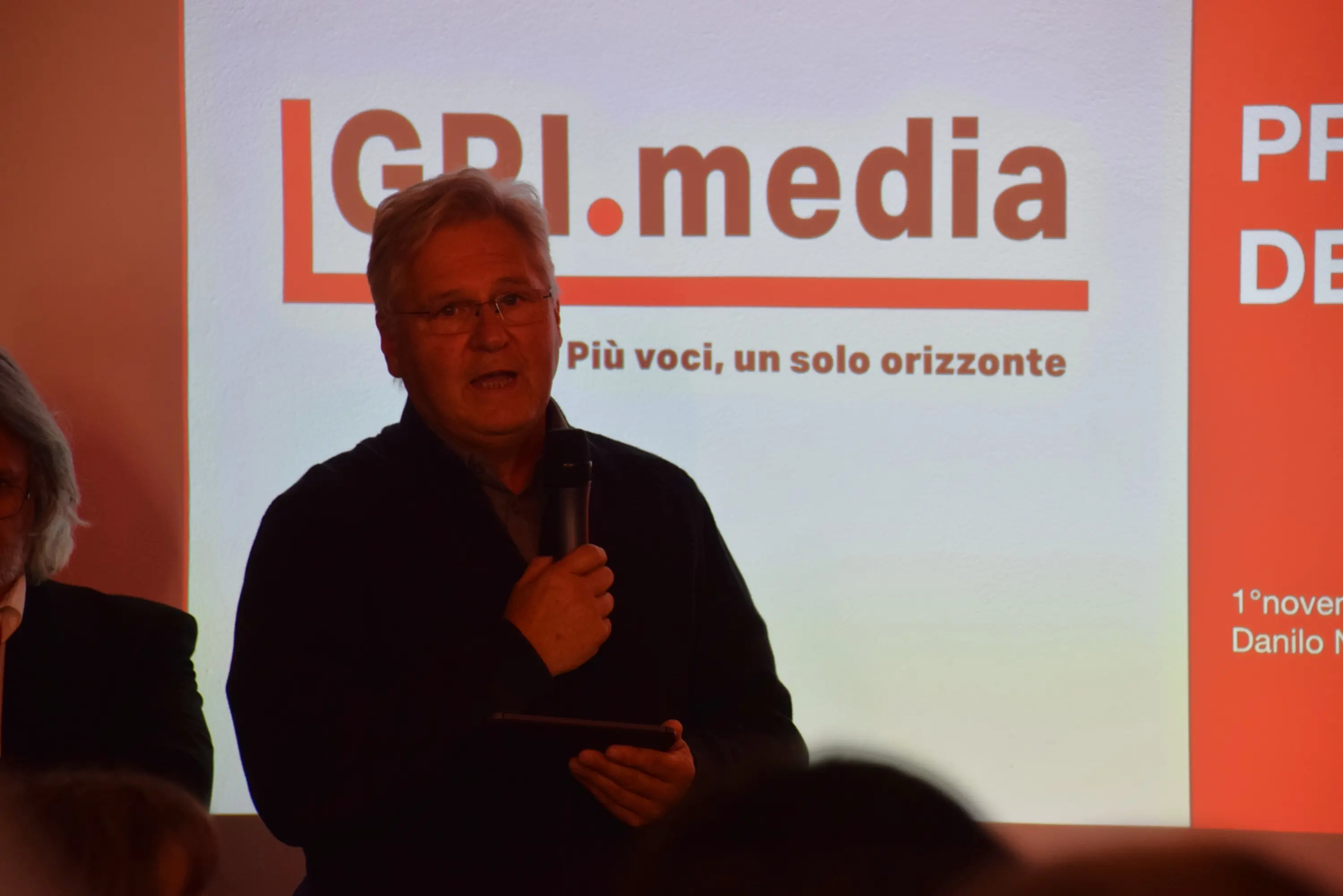 Un uomo con i capelli grigi parla al microfono durante un evento, con un logo di GPI.media sullo sfondo. Si tiene un tablet nella mano e sembra essere coinvolto in una presentazione.