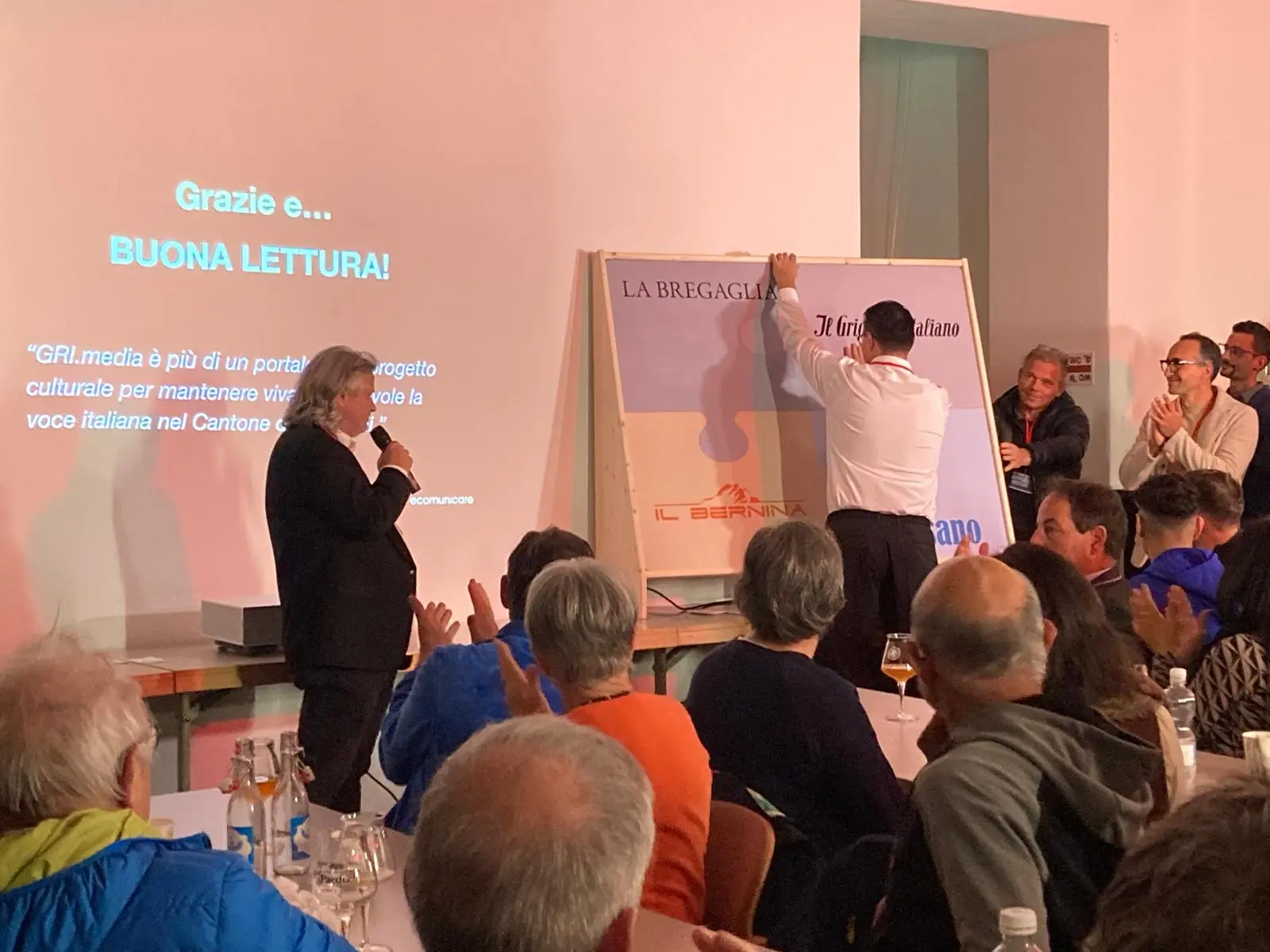 Un gruppo di persone applaude mentre un uomo scopre un grande poster su un leggìo. Sullo sfondo, un messaggio di ringraziamento e invito alla lettura. L'atmosfera è festosa e coinvolgente.