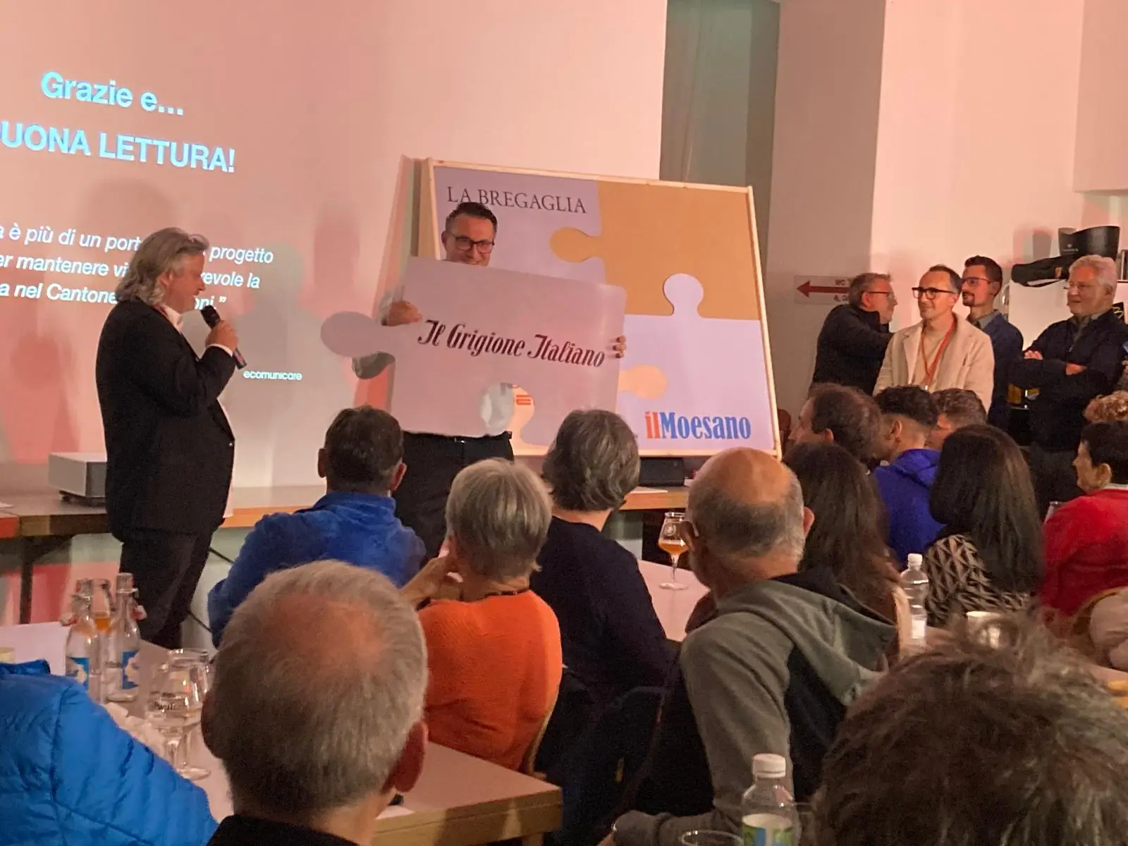 Un uomo tiene un cartello con il logo "Il Grigione Italiano" mentre parla a un pubblico numeroso in una sala. Altre persone ascoltano attentamente e sono visibilmente interessate all'evento.