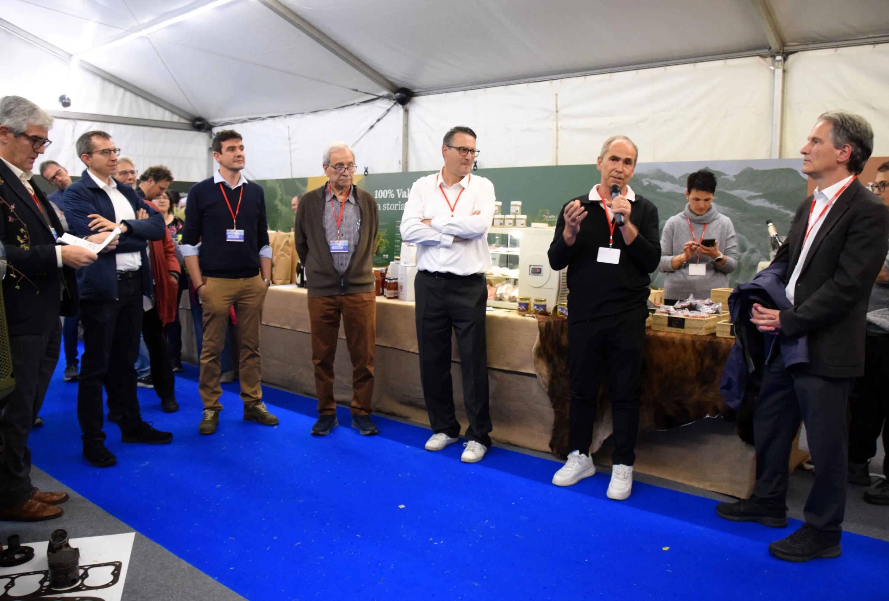 Un gruppo di persone riunite in un tendone, mentre un uomo al microfono parla. Sul tavolo dietro di lui ci sono vari prodotti alimentari e bevande. Alcuni partecipanti ascoltano attentamente, mentre altri sono impegnati con i loro dispositivi.