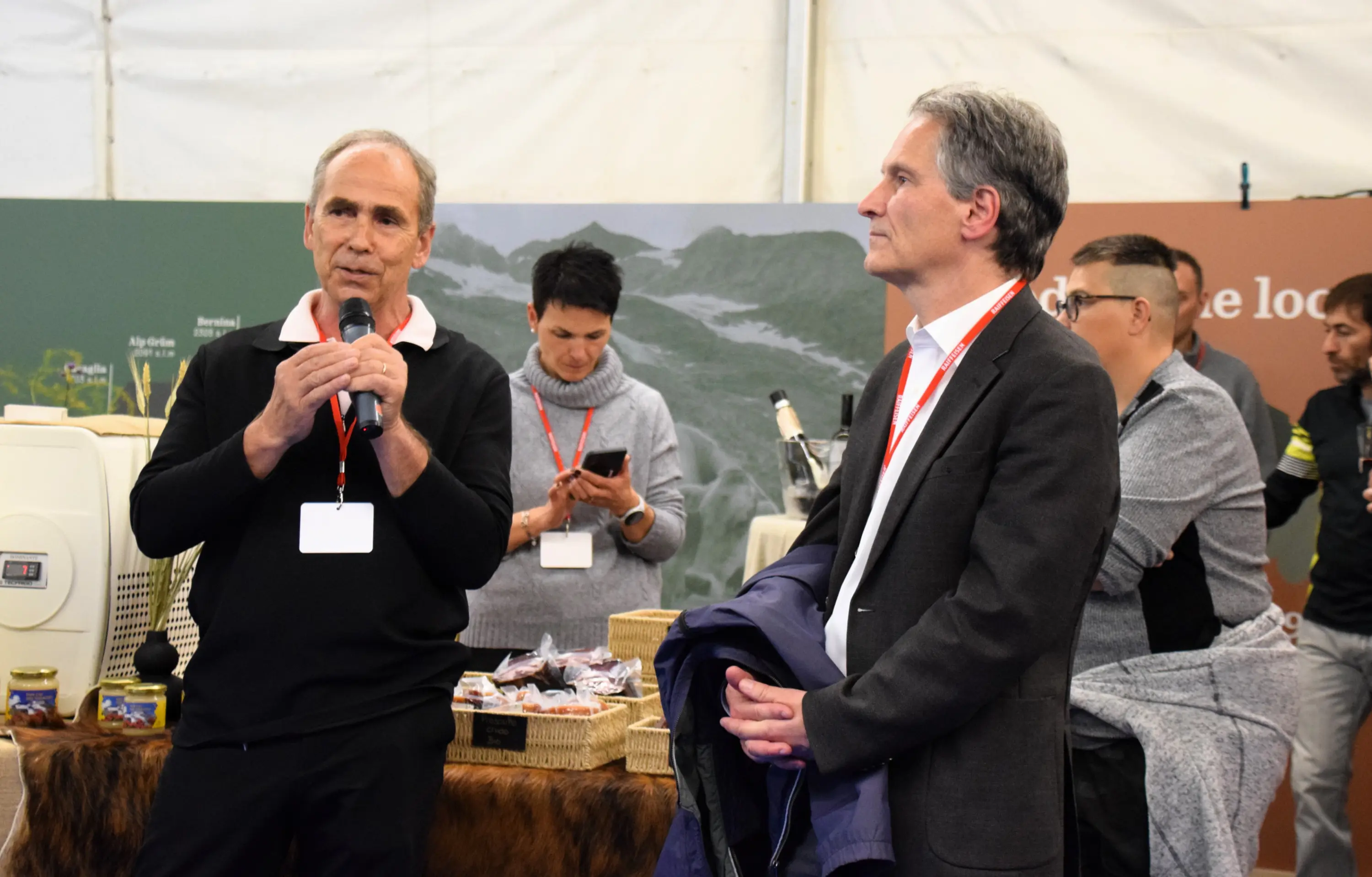 Un uomo parla al microfono mentre un altro lo ascolta attentamente. Sullo sfondo ci sono persone che interagiscono e diversi prodotti locali esposti.