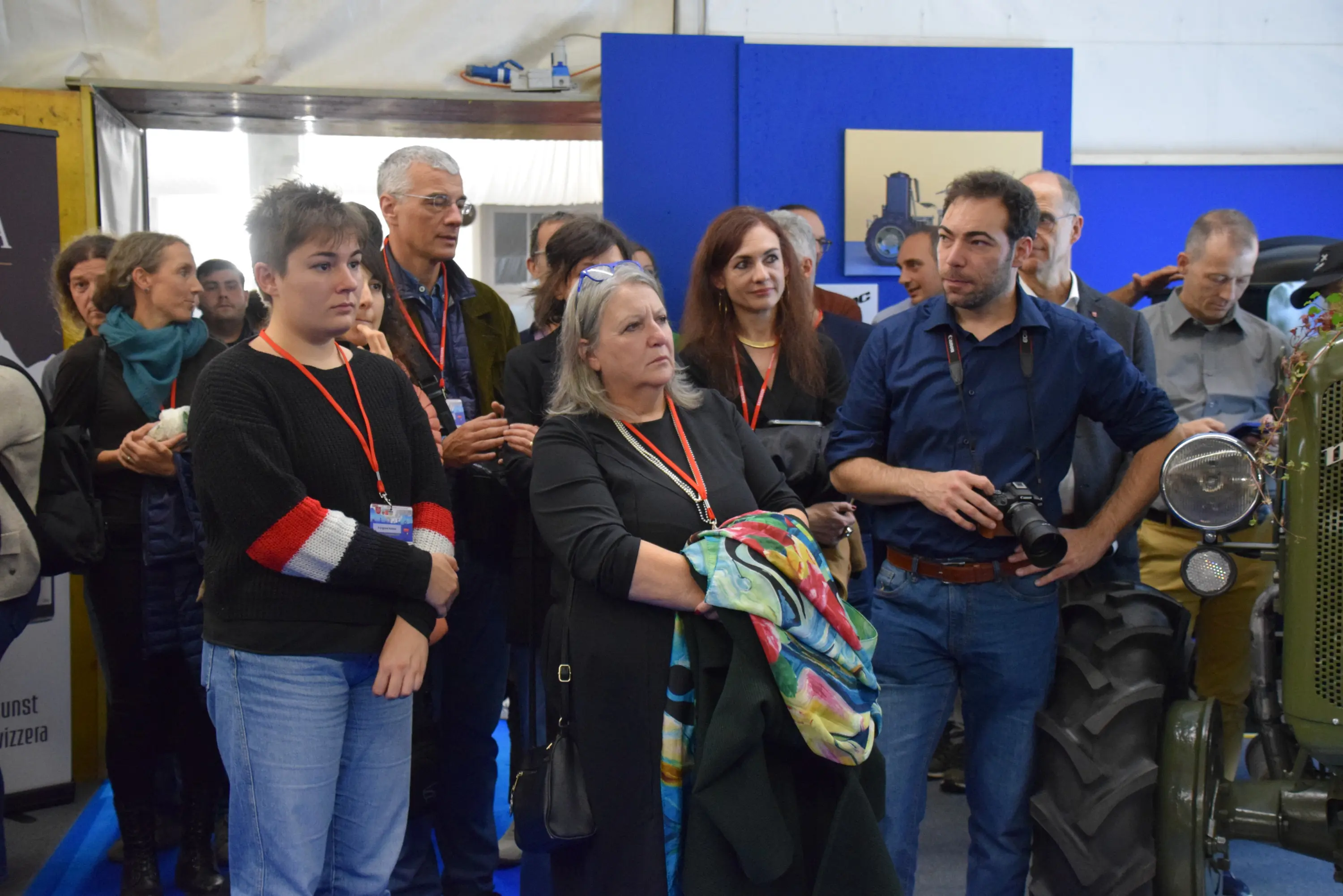 Un gruppo di persone in un evento, tutte concentrate su una presentazione. Alcuni indossano partecipazioni e altri hanno una macchina fotografica. Presenza di un trattore sullo sfondo.