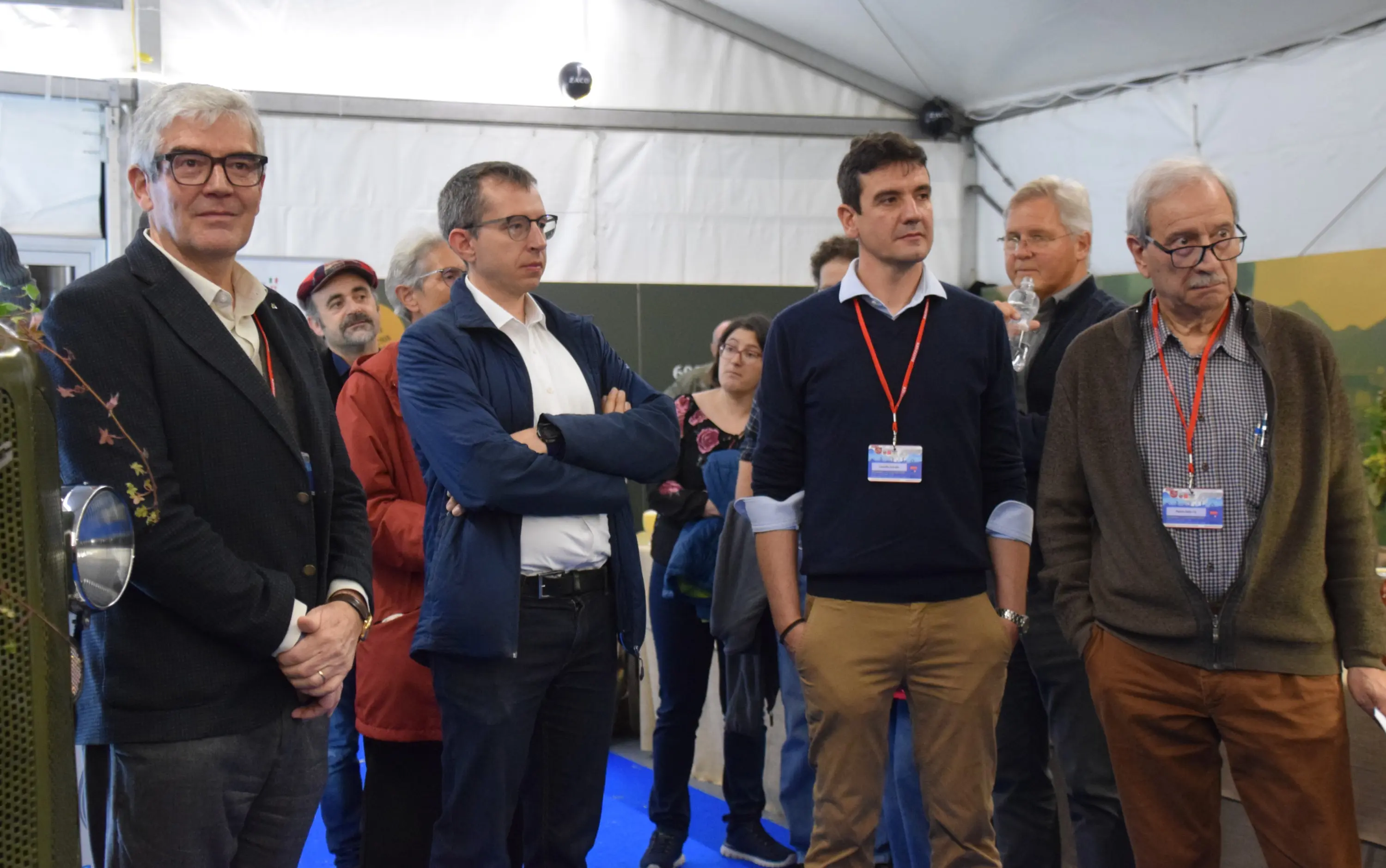 Gruppo di persone all'interno di una tenda, alcune in posa, altre osservano attentamente. Sono presenti espressioni di interesse e attenzione nei loro volti.