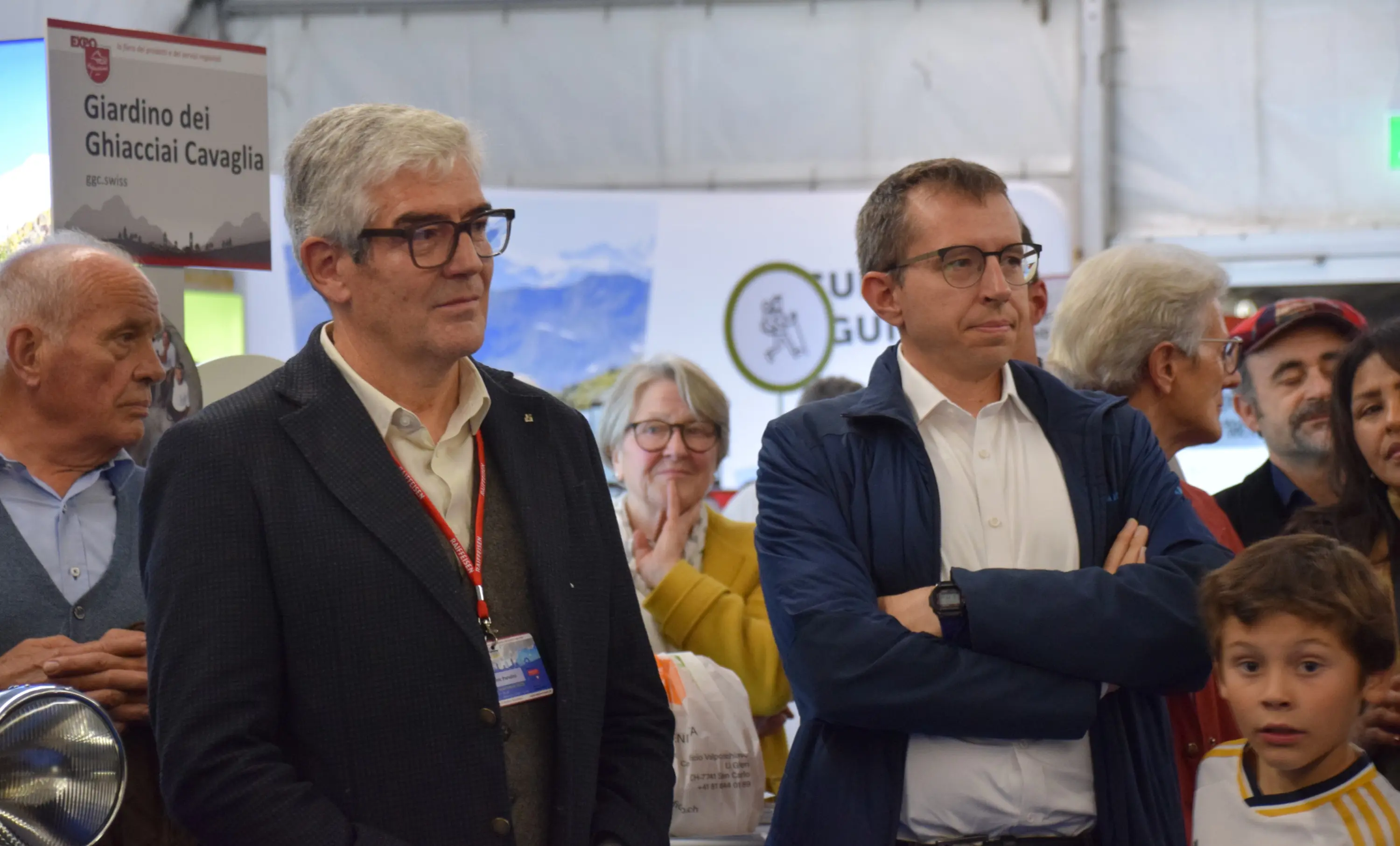 Due uomini in abiti formali osservano un evento, circondati da un pubblico di varie età. Alcuni partecipanti sembrano conversare mentre altri si concentrano sull'evento.