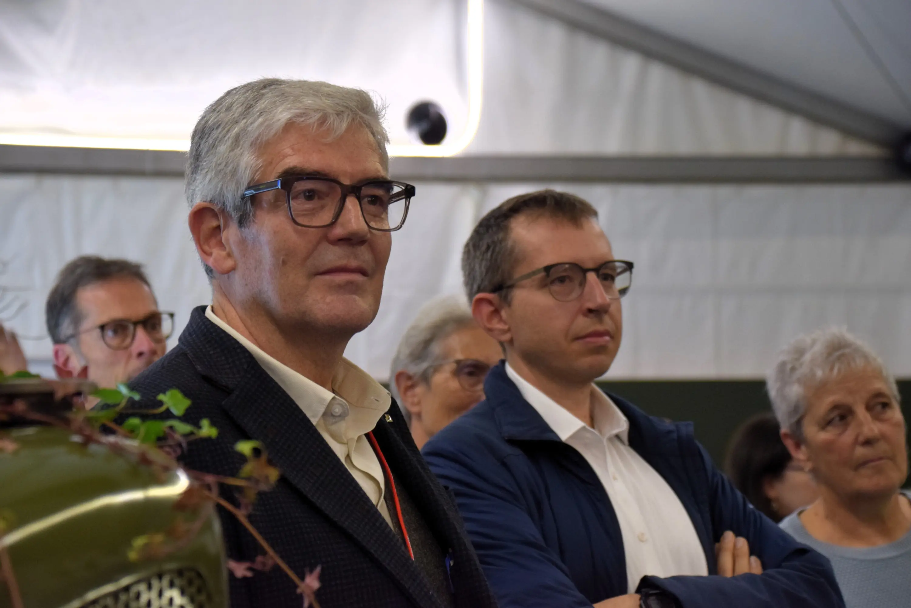 Un gruppo di persone ascolta attentamente durante un evento, con espressioni di interesse. Due uomini in primo piano indossano occhiali e sembrano riflettere su ciò che sta accadendo.