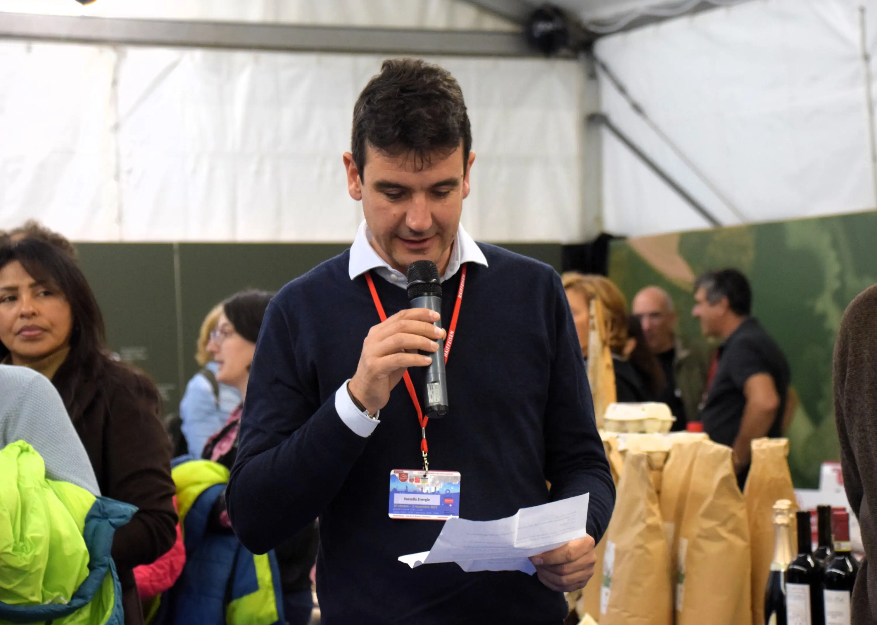 Un uomo in maglione scuro tiene un microfono mentre parla a un evento. Sullo sfondo, diverse persone ascoltano, con alcune che indossano giacche colorate.