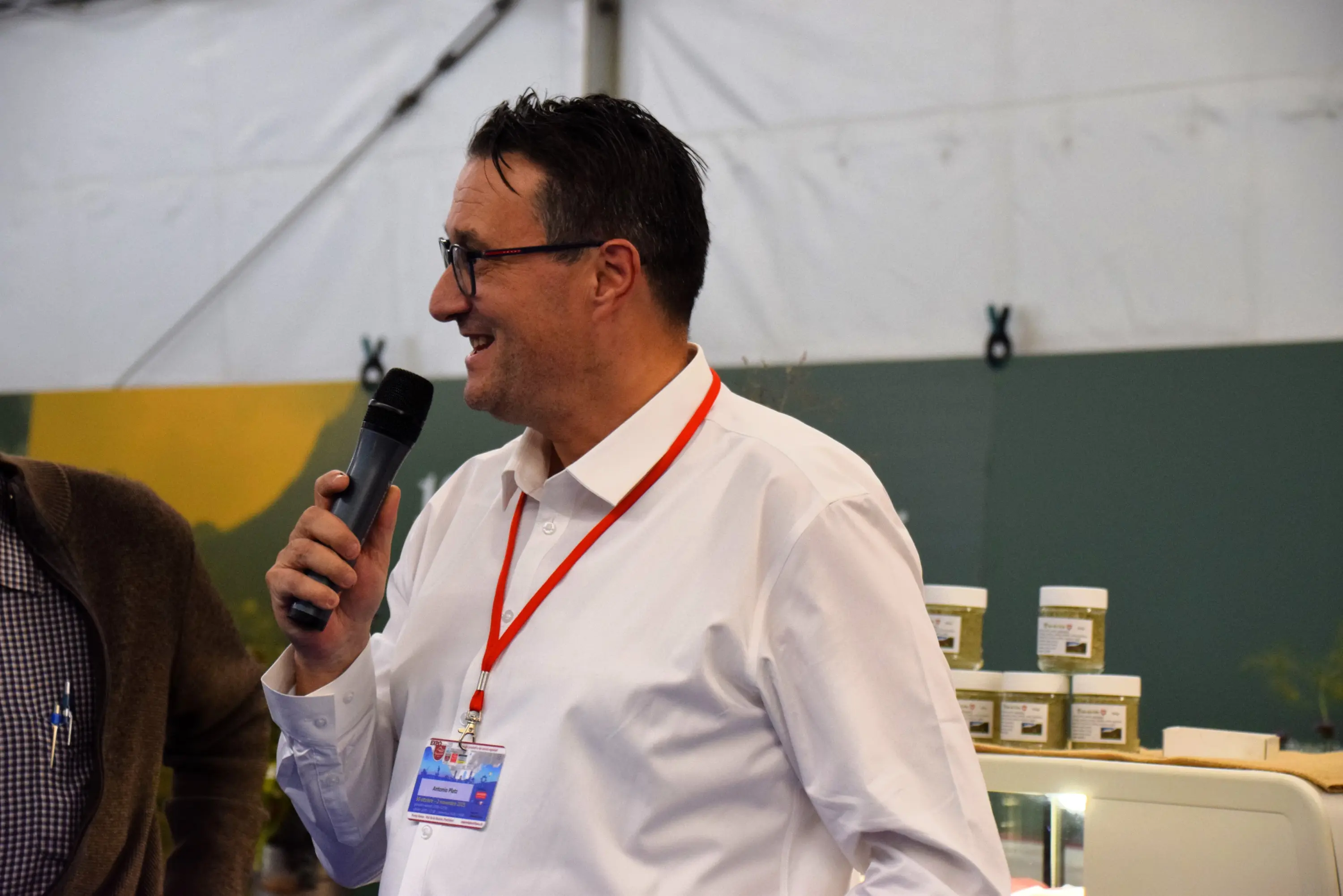 Un uomo con occhiali e camicia bianca tiene un microfono mentre parla a un pubblico, con un'espressione coinvolta. Sullo sfondo si vedono dei barattoli e una tenda che crea un'atmosfera di evento o fiera.