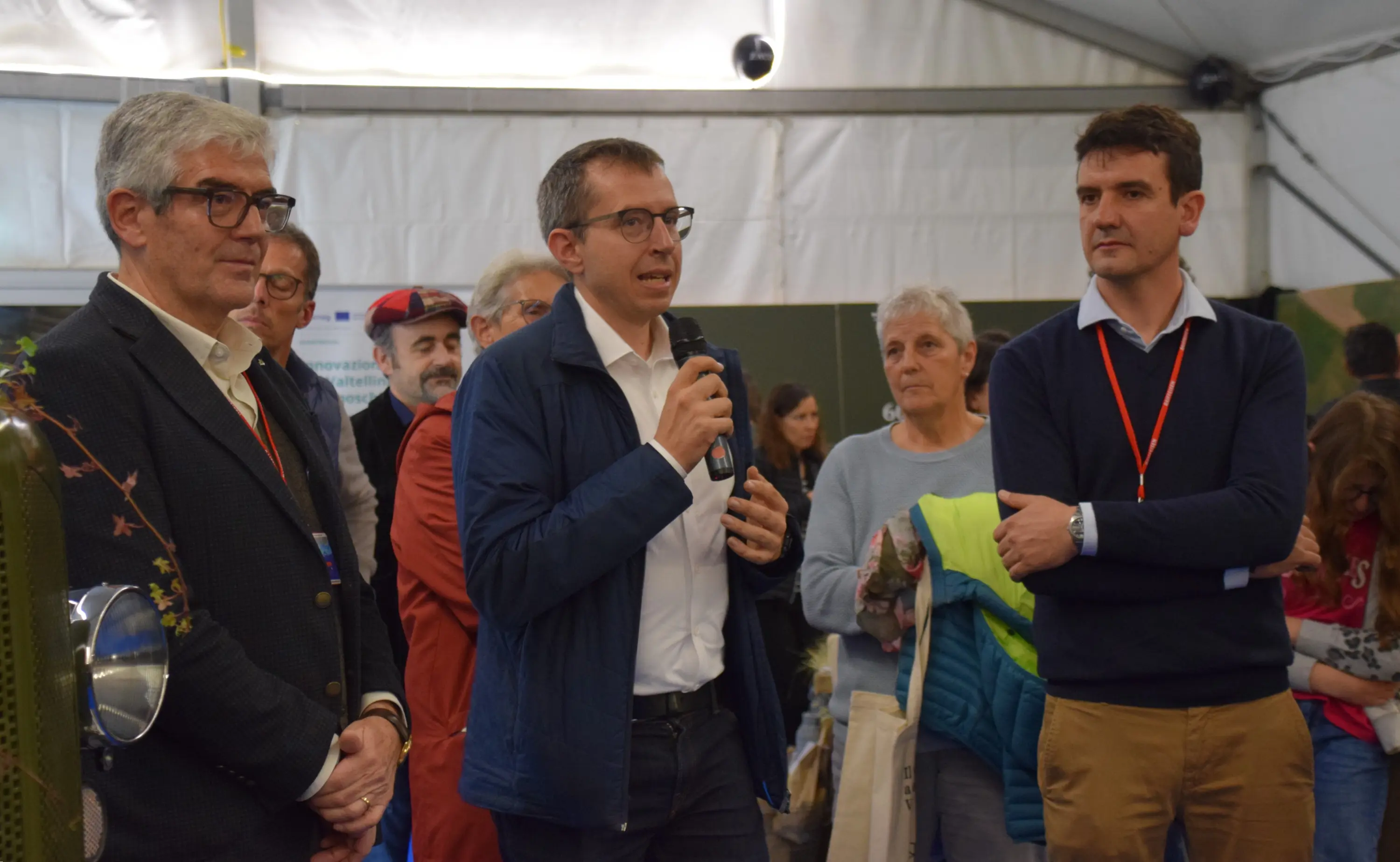 Tre uomini si trovano in un ambiente interno, probabilmente a un evento pubblico. Uno di loro parla al microfono, mentre gli altri ascoltano attentamente. Dietro di loro si possono vedere altre persone e un'atmosfera di scambio e discussione.