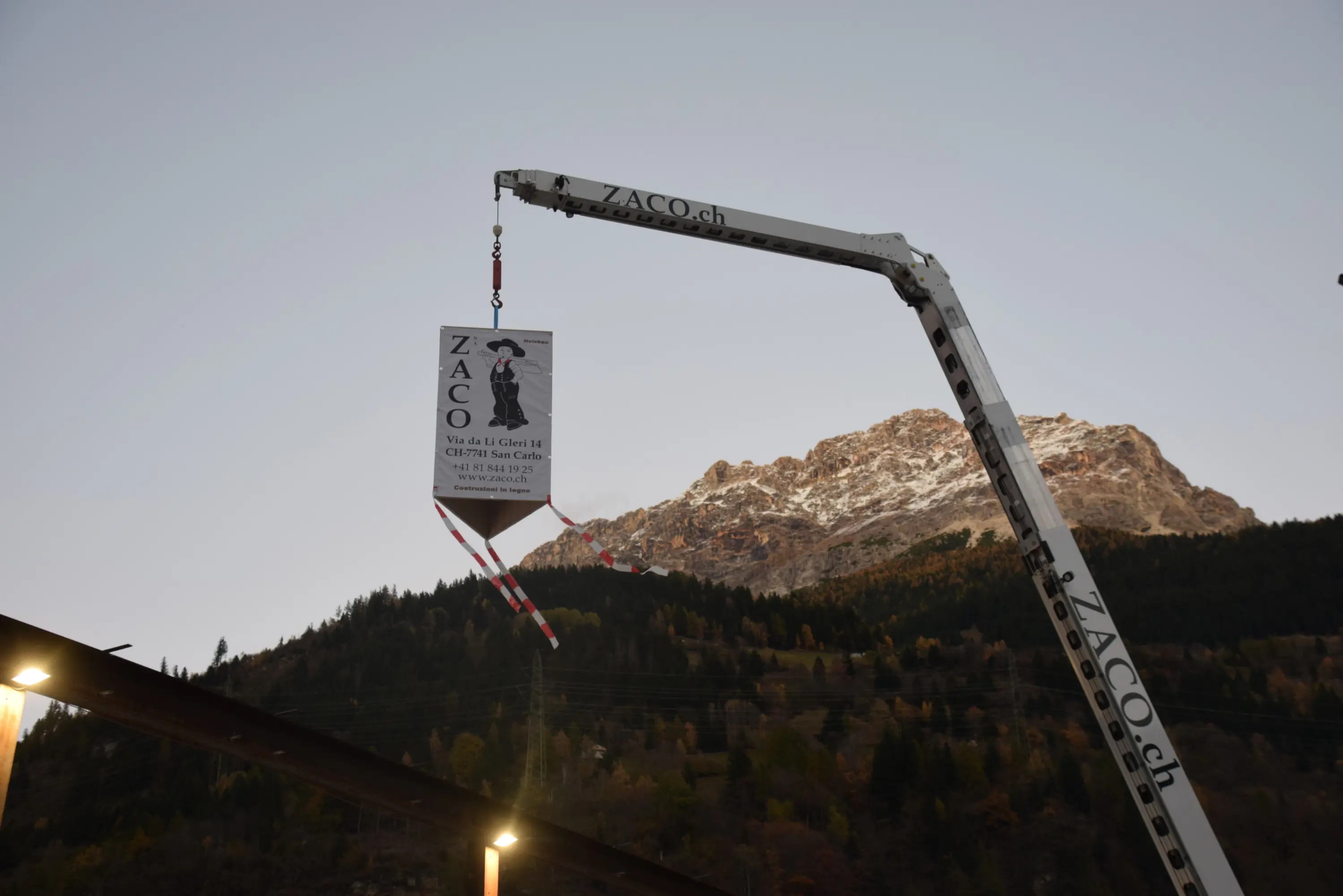 Un cartello di legno con il logo "ZACO" appeso a una gru, mentre sullo sfondo si intravede una montagna innevata al tramonto.