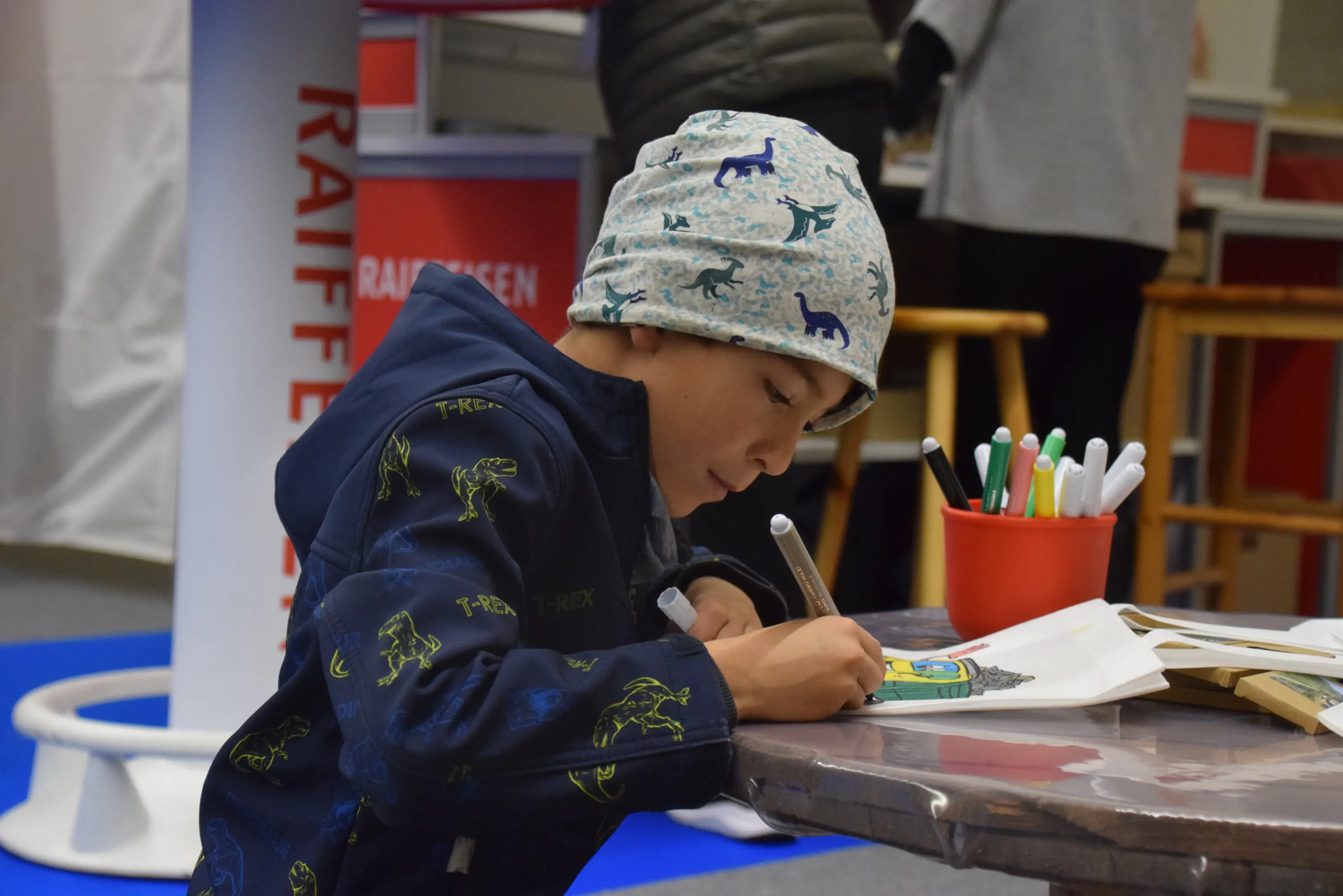 Un bambino seduto a un tavolo, mentre disegna con un pennarello. Indossa un cappello azzurro con disegni di animali e una giacca scura con motivi colorati. Sul tavolo ci sono colori e un libro da colorare.