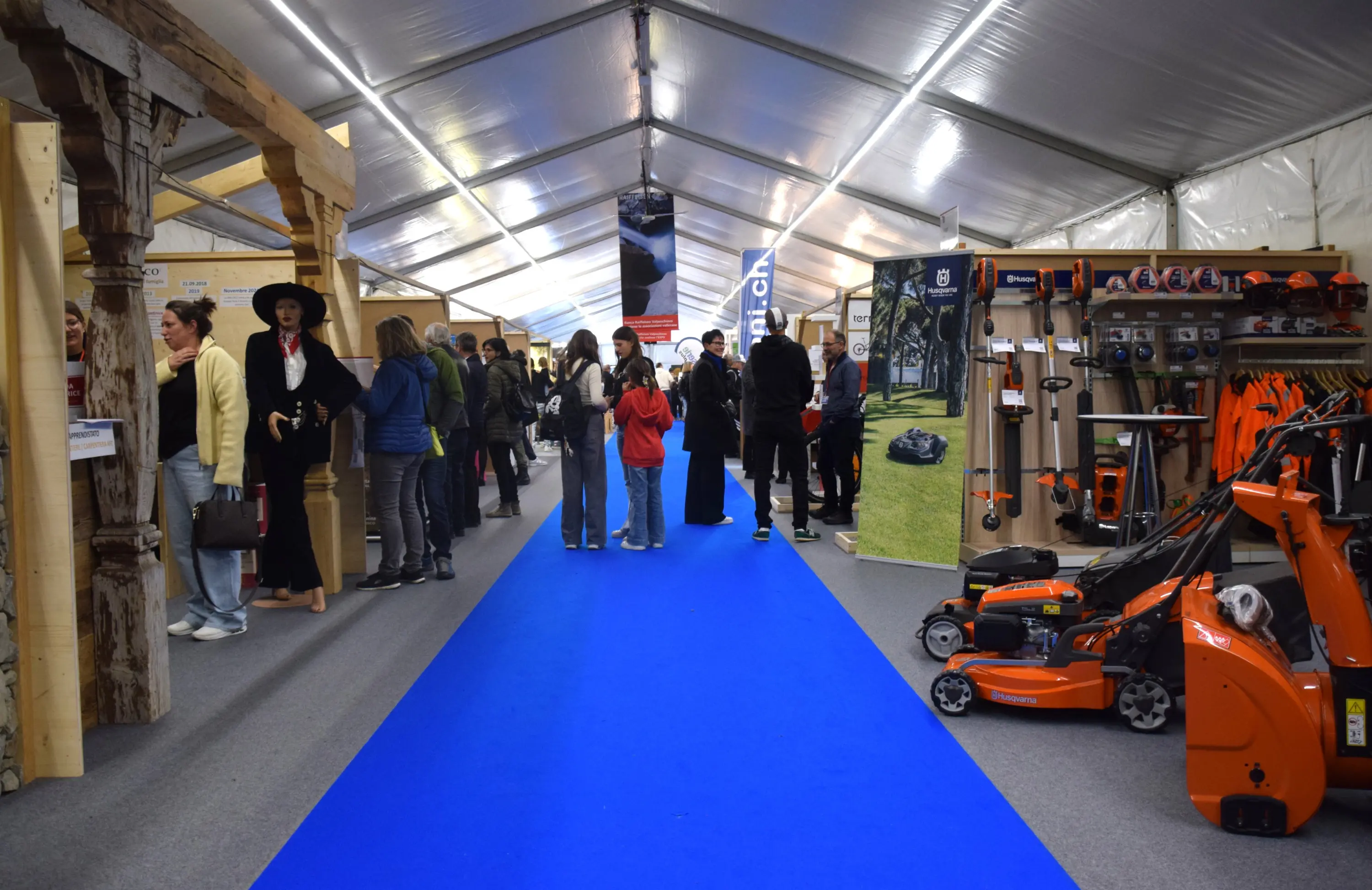 Una fiera all'interno di una tensostruttura con visitatori intenti a osservare vari stand. Il pavimento è coperto da un tappeto blu, mentre si possono vedere attrezzature per il giardinaggio e mobili esposti.
