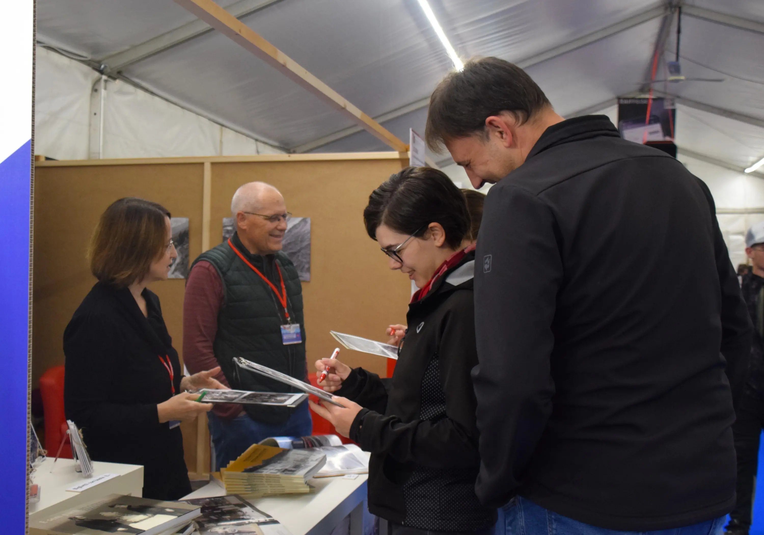 Persone all'interno di una tenda, con un tavolo dove alcuni visitatori stanno ricevendo autografi. Due persone sorridono mentre un uomo e una donna consegnano delle foto a un artista.