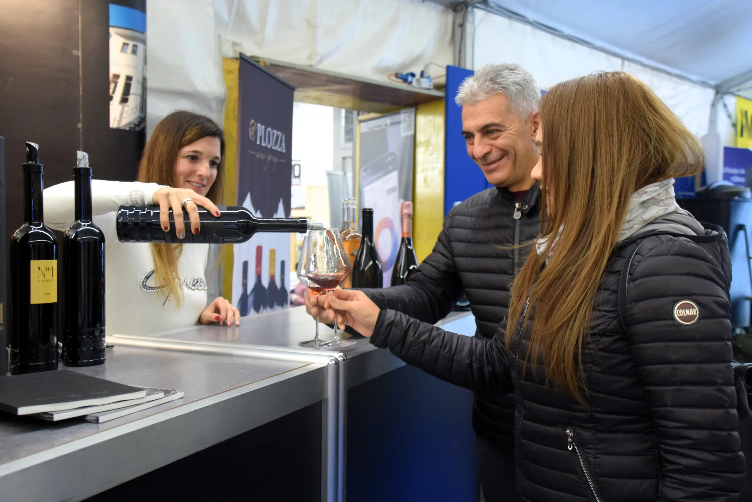 Una sommelier versa del vino in un bicchiere per una coppia, mentre sono in un evento con vari vini esposti. L'atmosfera è festosa e conviviale.