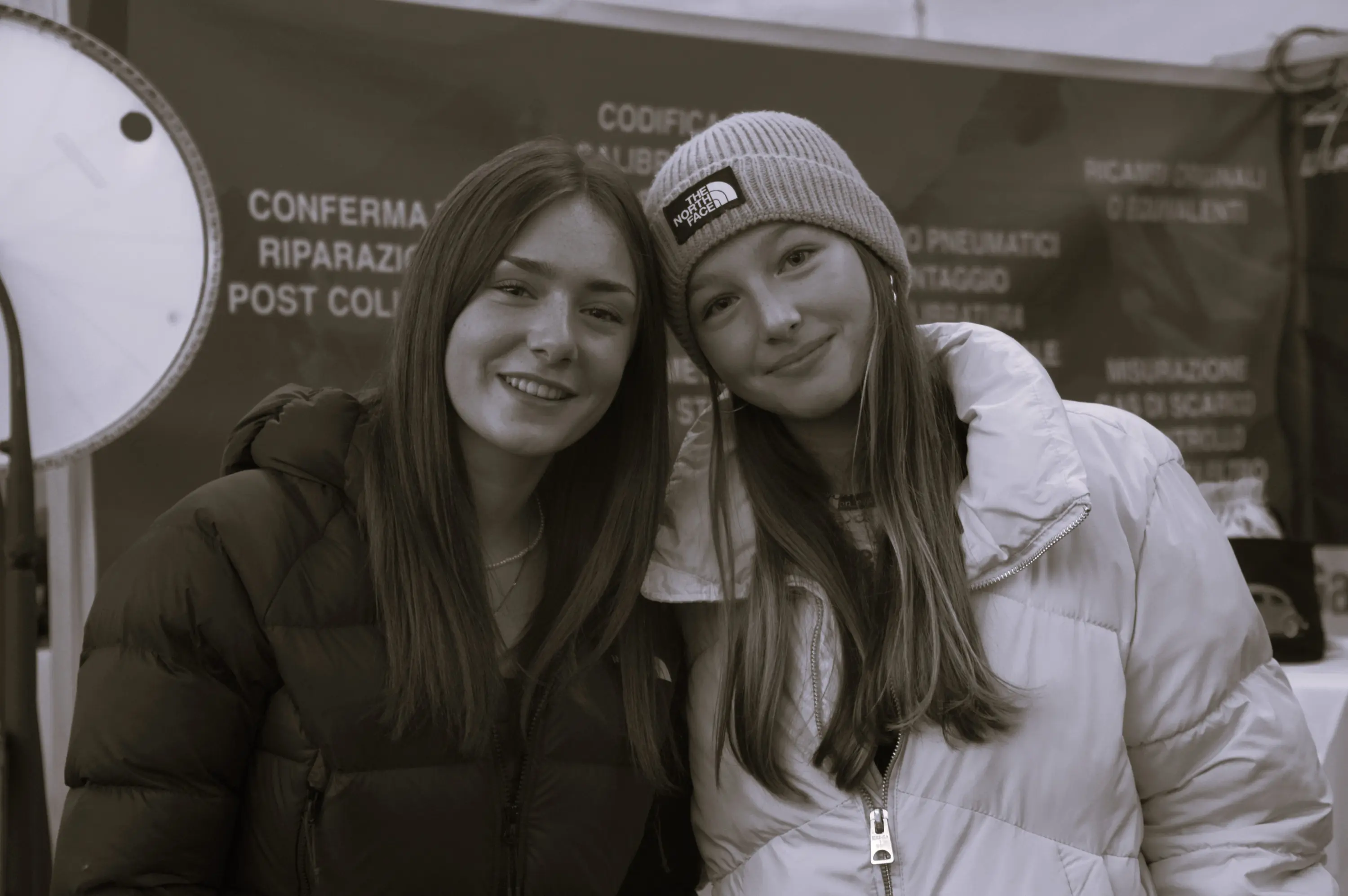Due ragazze sorridenti posano insieme, indossando giacche invernali e berretti, con uno sfondo di un mercato o evento.