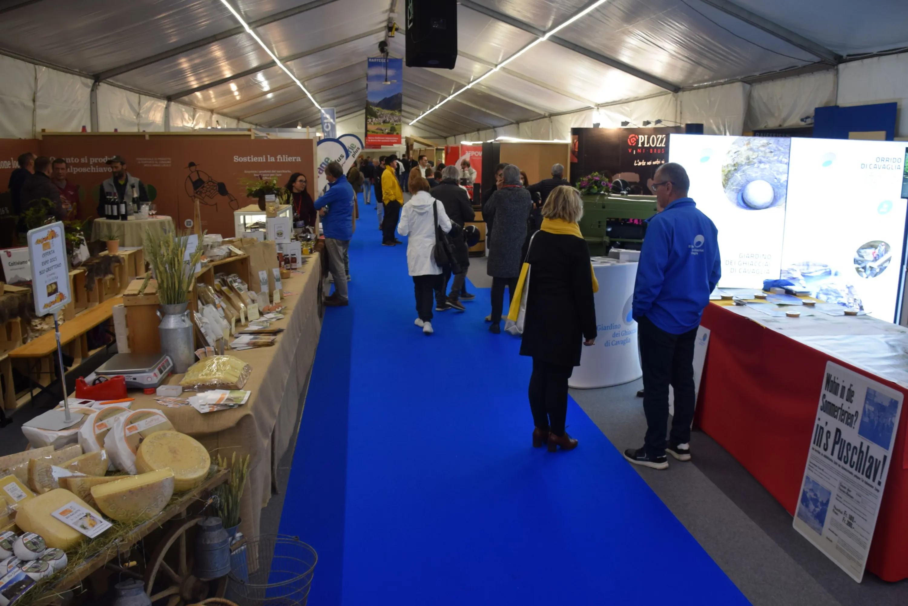 Un'area espositiva coperta con tunnel, con stand di prodotti locali, come formaggi e cereali. Persone passeggiano lungo un corridoio blu, mentre alcuni visitatori si fermano per osservare le offerte. In fondo, stand di diverse aziende e diverse presentazioni visive.