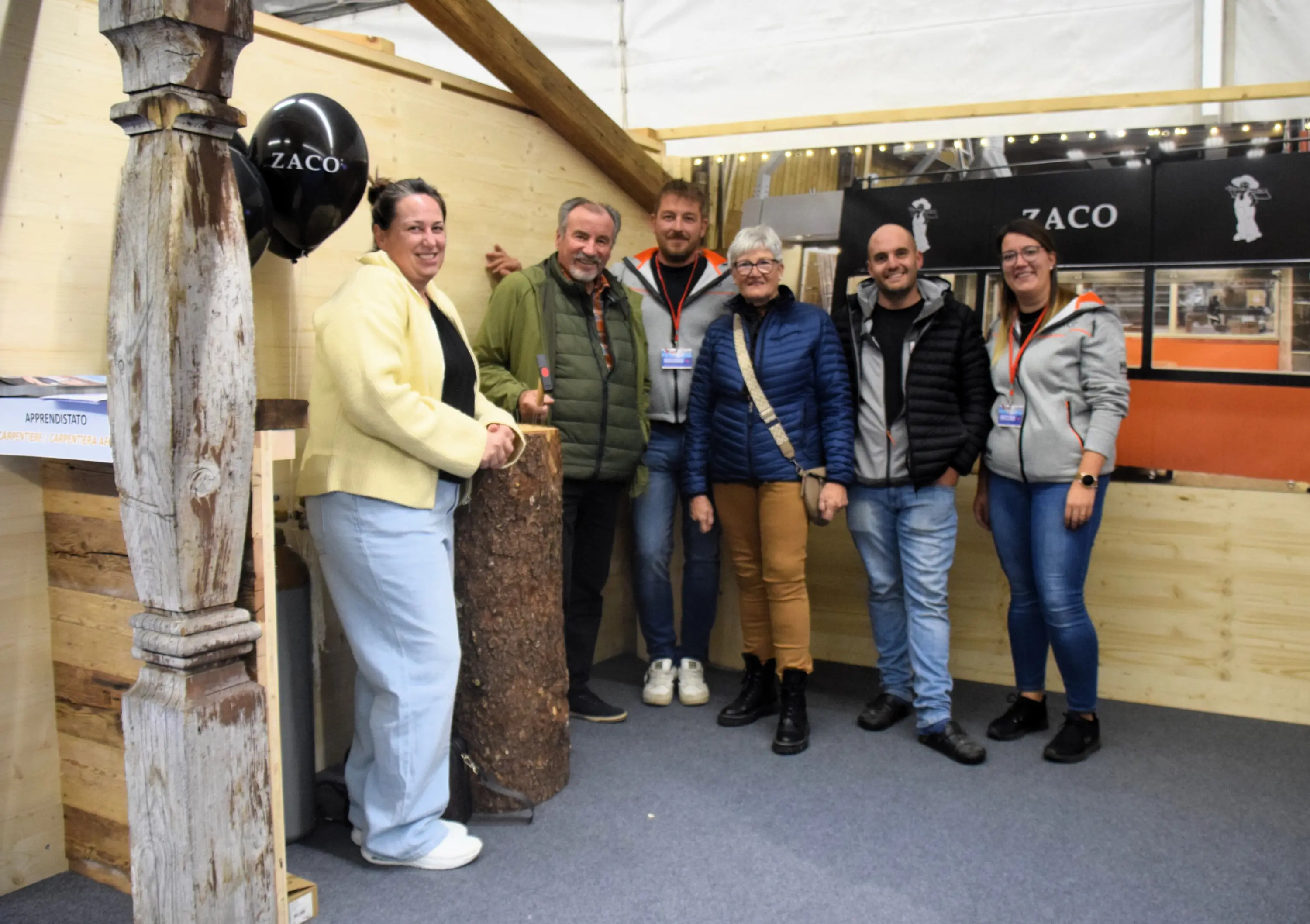 Un gruppo di sei persone sorridenti, vestite in modo informale, posano insieme in uno stand fieristico. Sullo sfondo si vedono materiali di legno e un pallone con il logo "Zaco".