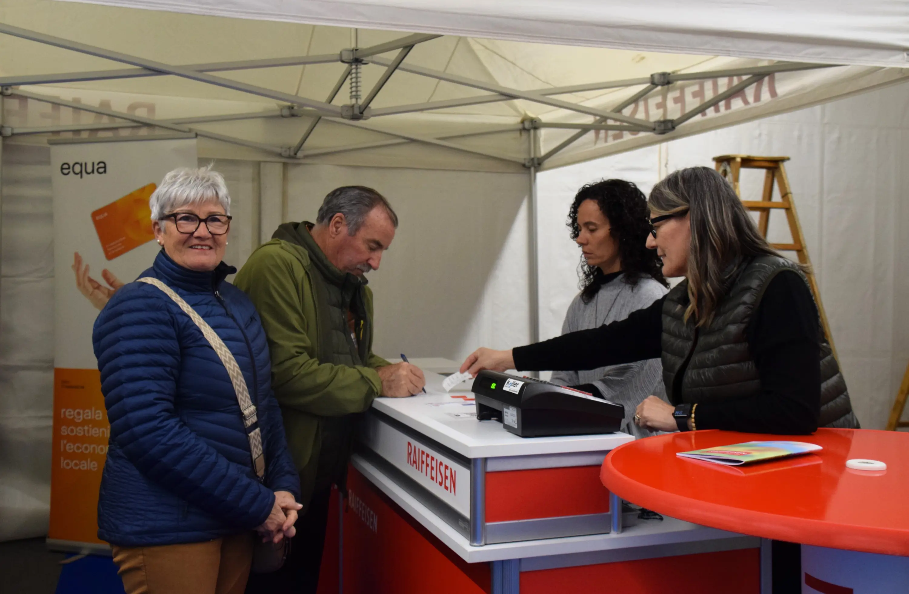 Una donna sorridente in primo piano, con occhiali e giacca blu. Dietro di lei, due persone stanno compilando un modulo presso uno stand. Un'altra persona li osserva mentre scrivono. Insegne visibili con il logo di Raiffeisen sul bancone.