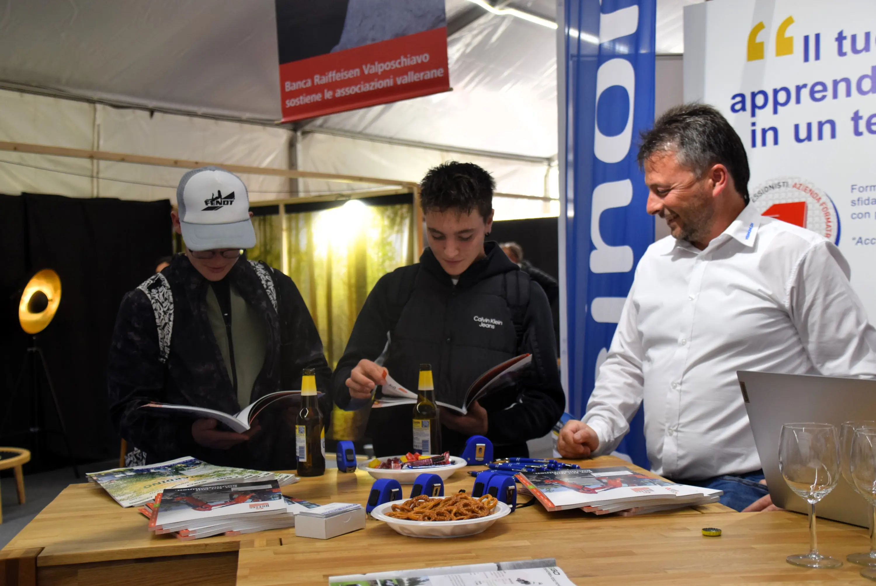 Due ragazzi esaminano dei cataloghi in una fiera, mentre un uomo adulto sorride e li osserva. Sul tavolo ci sono alcune bottiglie, snack e materiale promozionale. Sullo sfondo si intravedono pannelli pubblicitari.