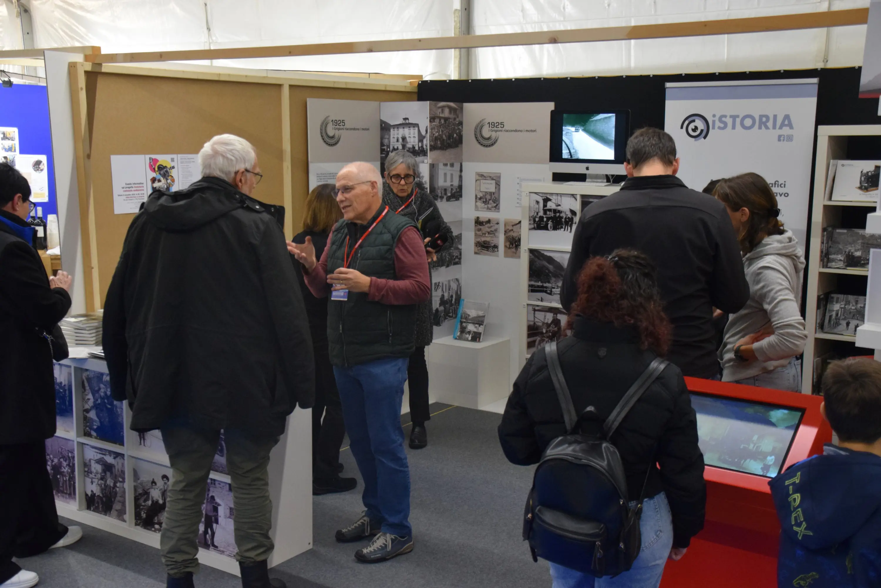 Una fiera con diverse persone che interagiscono. Un anziano sta parlando con un uomo, mentre altri visitatori osservano mostre fotografiche e interagiscono con un display digitale. Sullo sfondo si vedono pannelli informativi e fotografie storiche.