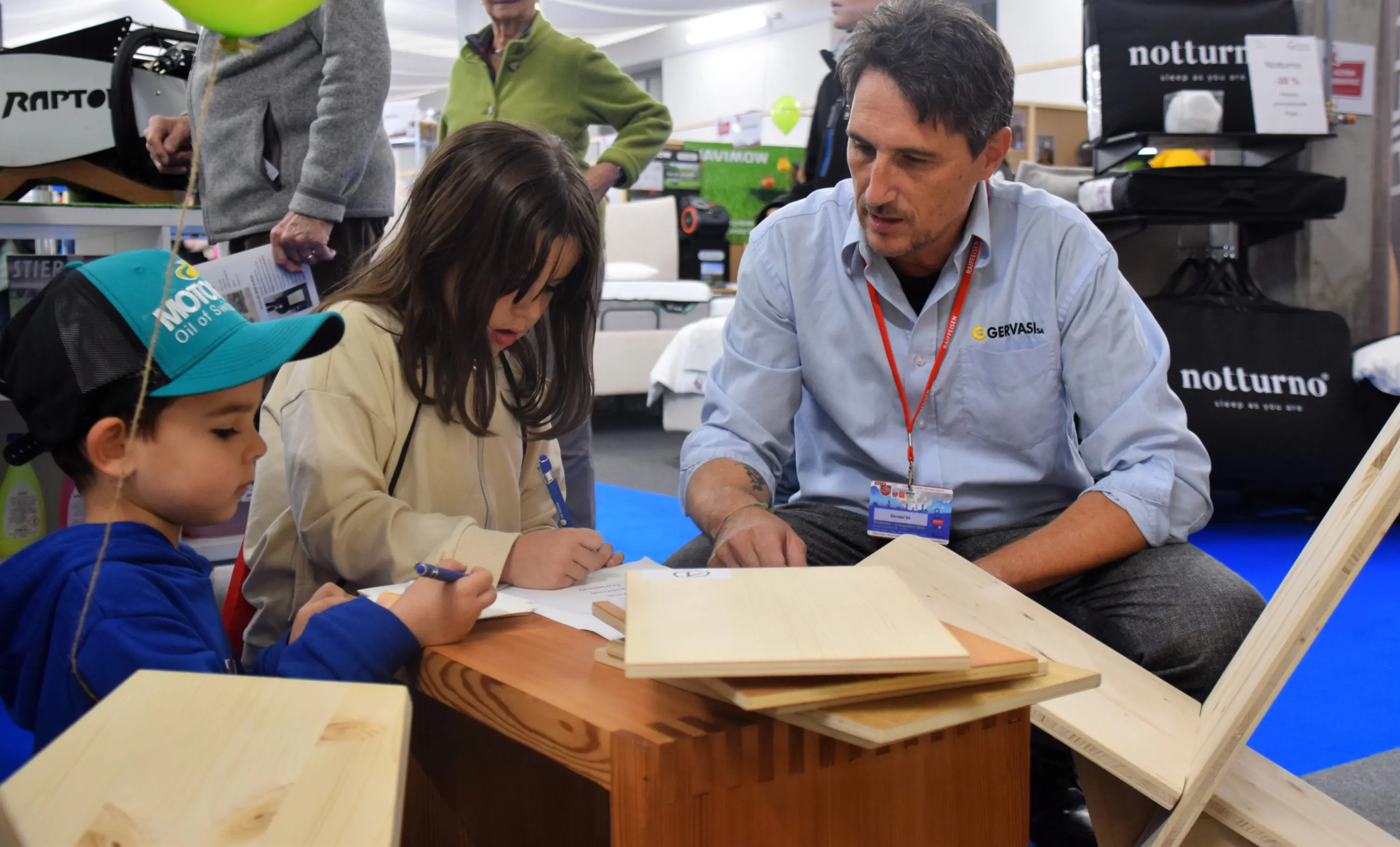 Un uomo parla con due bambini mentre si trovano attorno a un tavolo di legno. I bambini disegnano su un foglio, mentre l'uomo li osserva e interagisce con loro. Il contesto sembra essere un evento o una fiera.