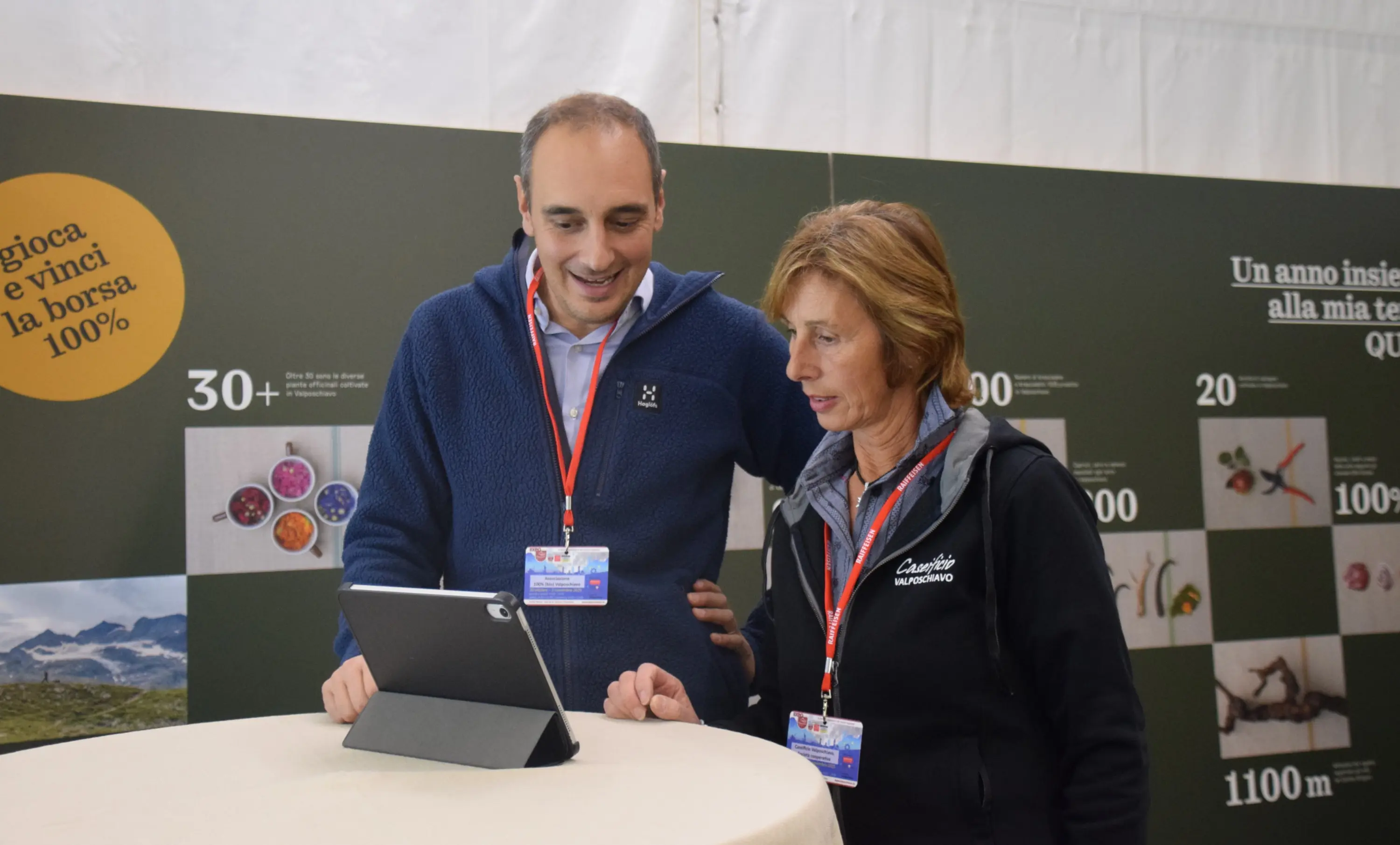 Due persone, un uomo e una donna, osservano un tablet mentre discutono, davanti a un pannello informativo con immagini e dati. Entrambi indossano giacche casual e hanno un distintivo con una cordicella.
