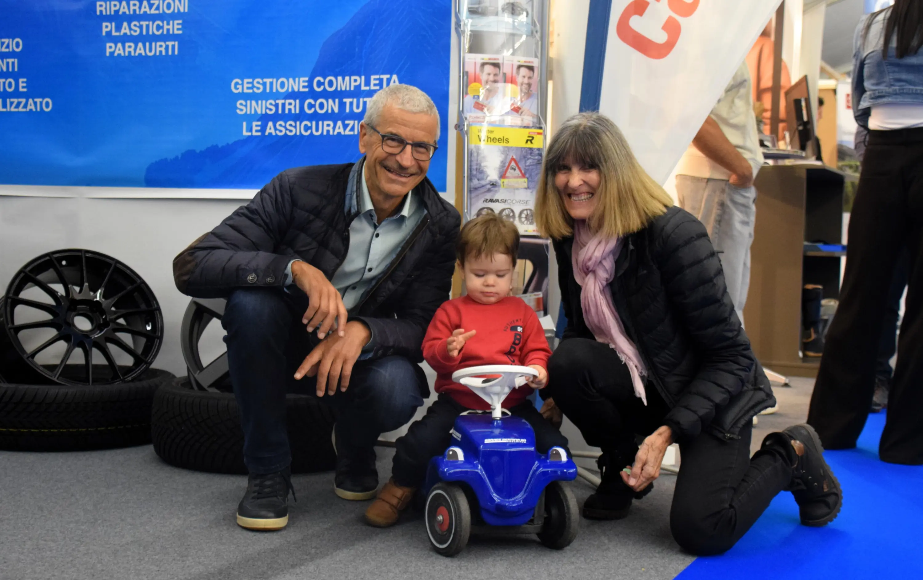 Una coppia sorride accanto a un bambino che gioca su un piccolo veicolo blu. Sullo sfondo, si vedono pneumatici e un banner con informazioni aziendali.