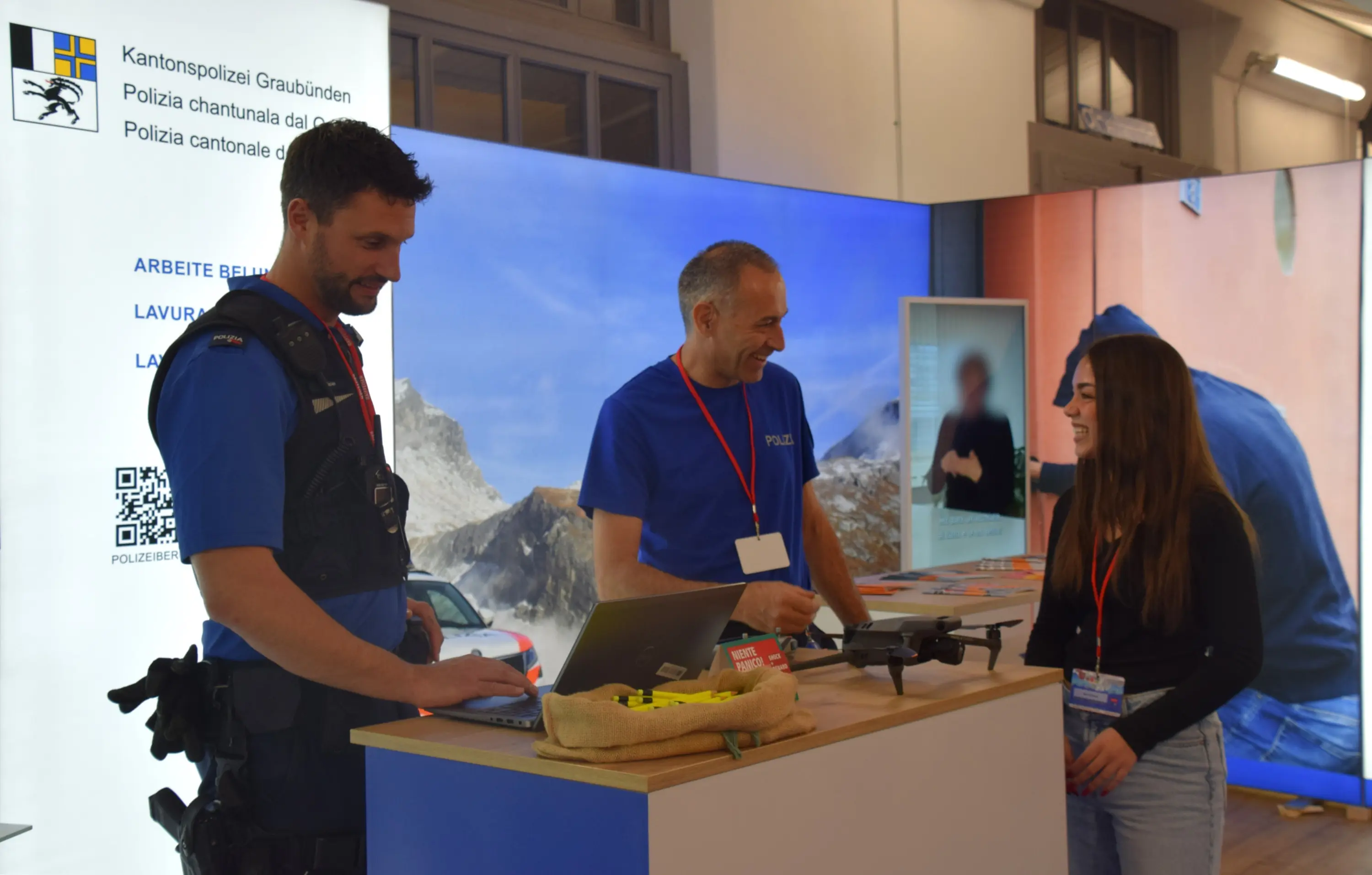 Due agenti di polizia sorridono mentre parlano con una giovane donna presso uno stand, con un computer portatile e un drone sul tavolo. Sullo sfondo, un grande schermo mostra immagini e informazioni relative alla polizia.