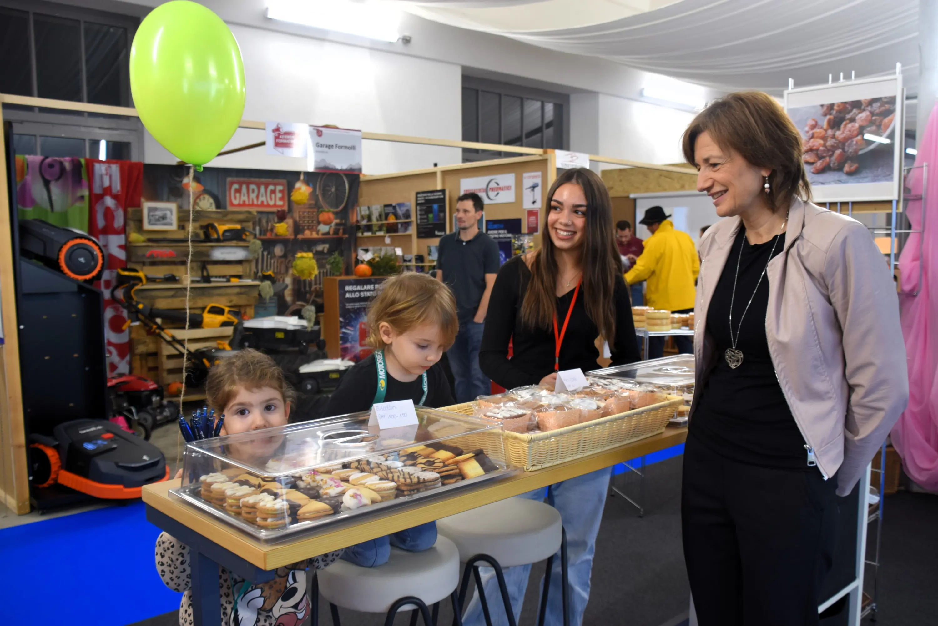 Due bambini osservano una vetrina di dolci di fronte a due donne sorridenti in un evento o fiera. Una donna, elegantemente vestita, sembra conversare con i bambini. Sullo sfondo si intravede un'area espositiva.